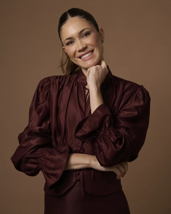 A young person with curly hair smiles at the camera while sitting in front of a laptop against a dark background.
