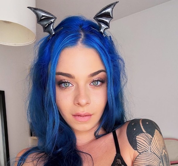 Woman in sunglasses poses against vibrant red background, wearing a beige blazer and black top. Sunlight casts dynamic shadows, creating a stylish, confident mood.