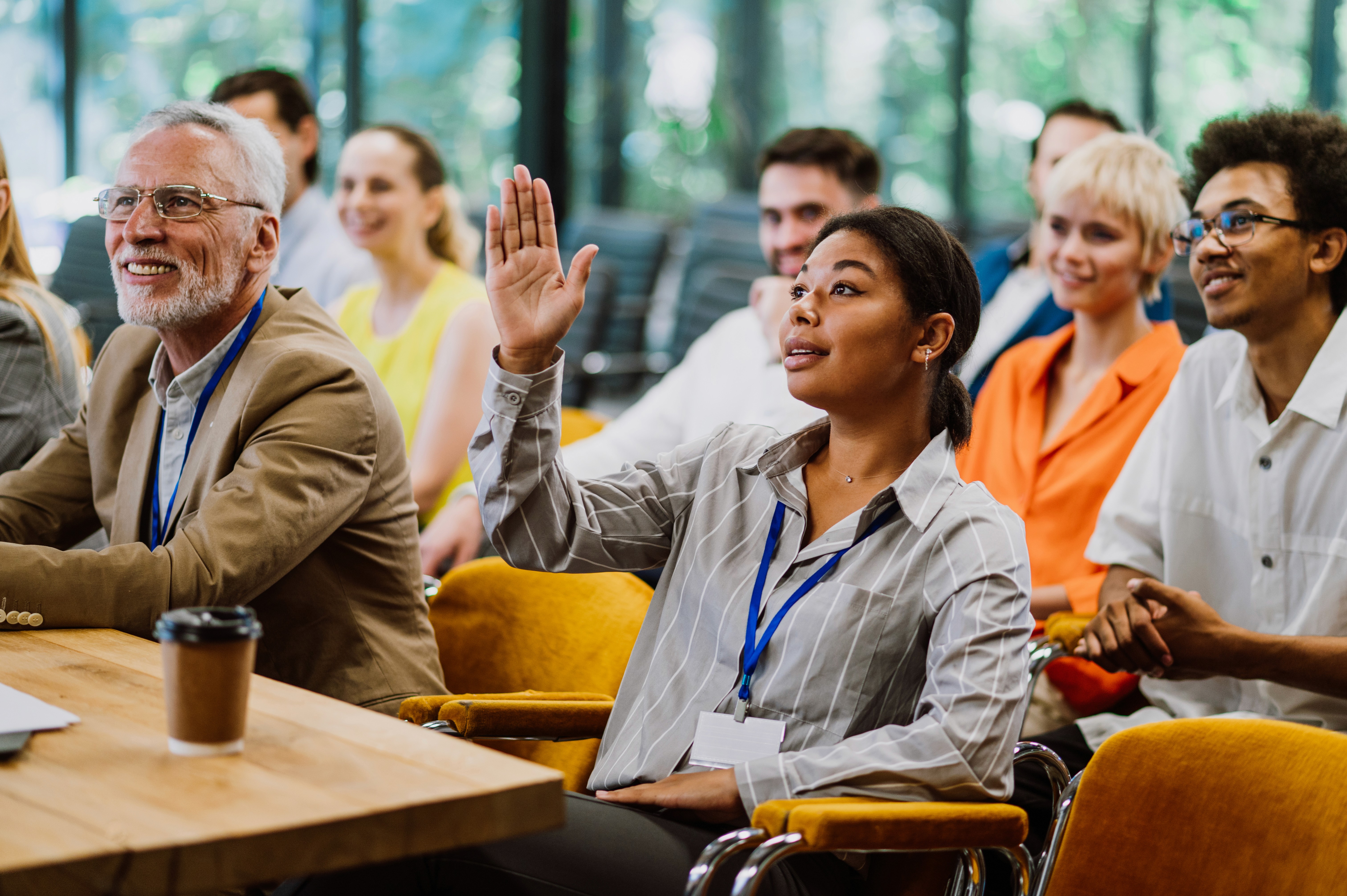 Man wearing an olive green jacket and black shirt, sitting and smiling in front of a light wooden background.