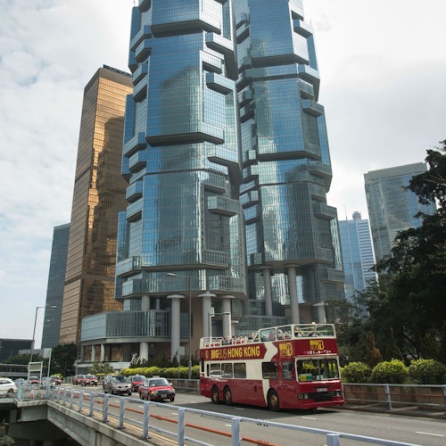 在城市环境中，一辆红色双层巴士行驶在现代玻璃摩天大楼和其他车辆之间的道路上。