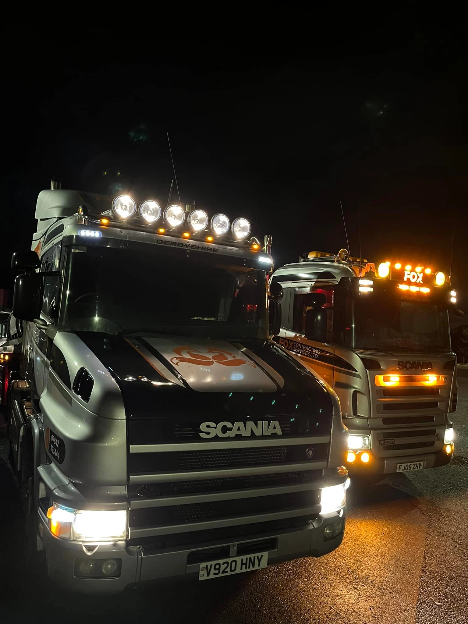 A large industrial facility with gray metal siding and a tall smokestack is in the background, while a white semi-truck towing a cylindrical tanker is parked in the foreground.