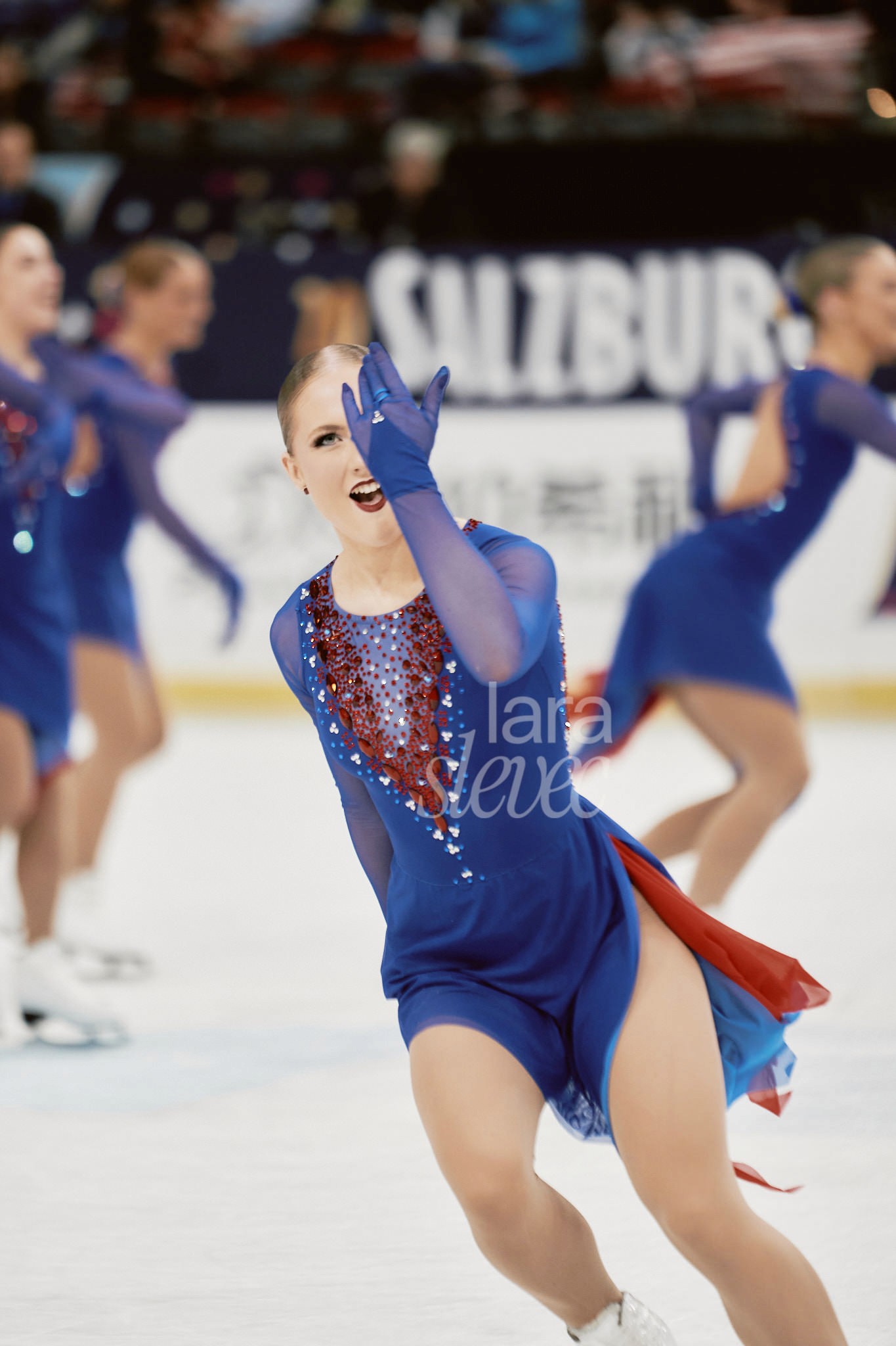 Synchroneiskunstläuferin, die eine Hand vor dem Auge hat, dabei aber in die Kamera schaut. Team Icicles. ISU Synchronized Skating Championships 2026 in Salzburg. Fotografiert von Lara Slevec.