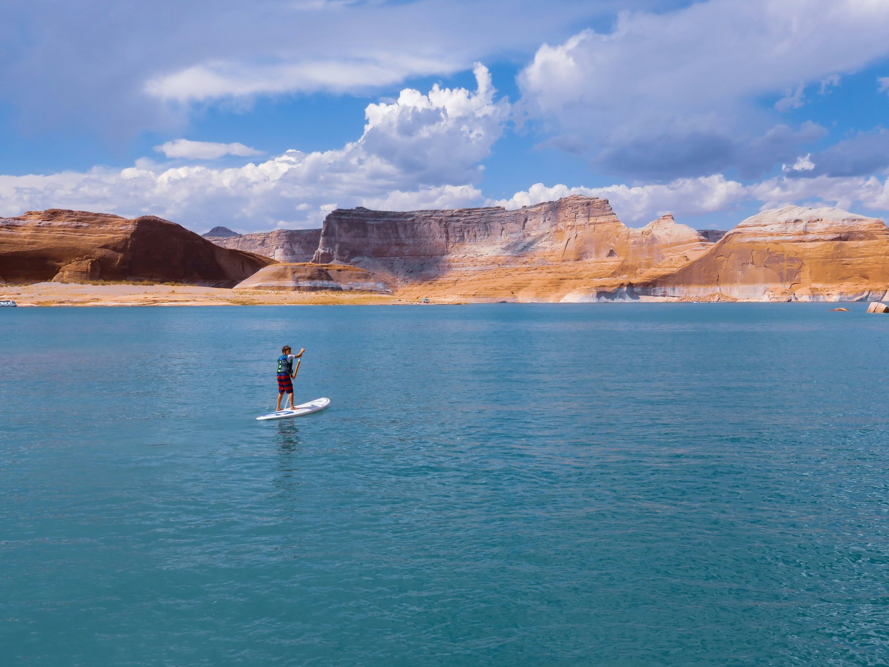 A paddleboarder crossing turquoise water on Lake Powell, with a pale band of bare rock along the shoreline marking the drop in water level.