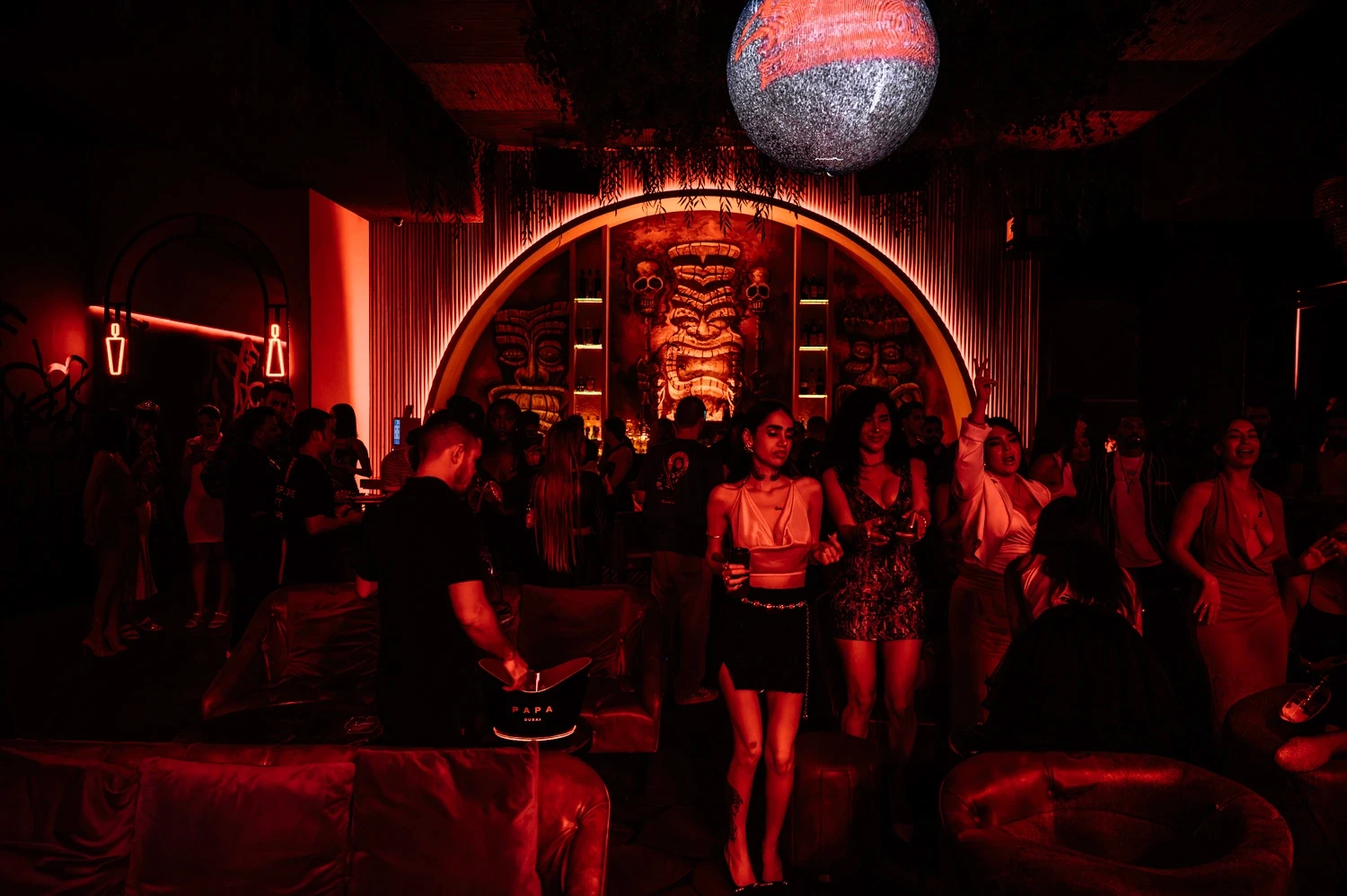 Red-lit lounge at PAPA Dubai with leather sofas, a disco ball and a tiki-themed bar under a glowing arch