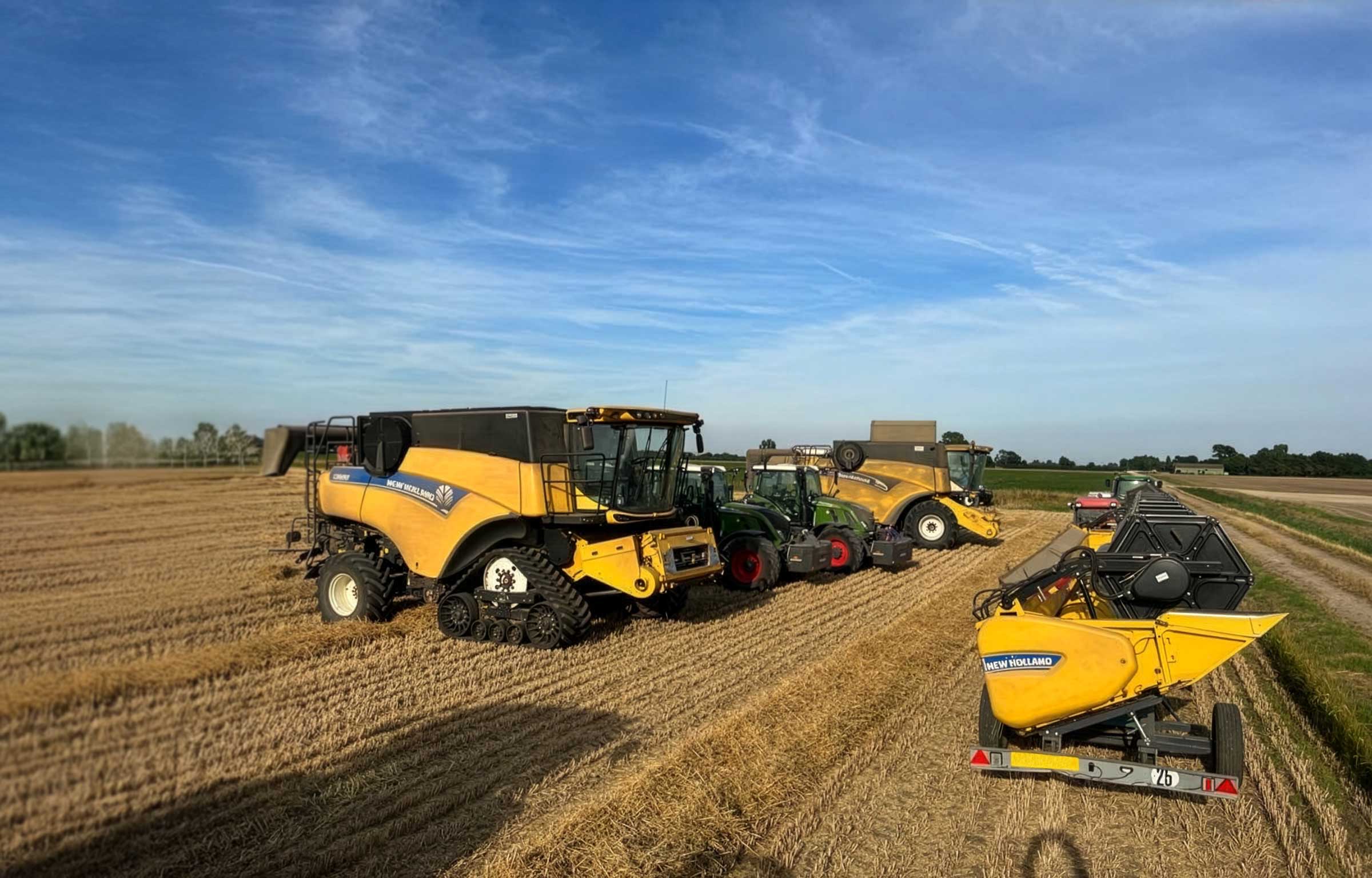 Fleet of yellow and green New Holland and Fendt farm machinery including combine harvesters and tractors in harvested field under blue sky