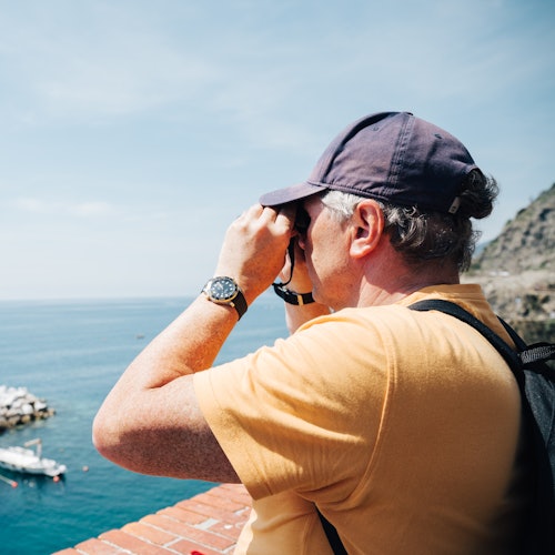 Osoba v žlutém tričku a kšiltovce používá dalekohled, aby se podívala na oceán z balkonu s cihlovým zábradlím.