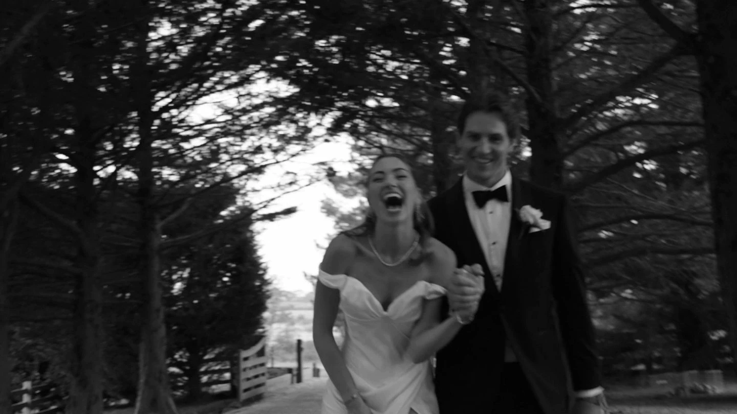 A joyful couple in formal attire walks hand in hand down a tree-lined path, surrounded by tall trees, as they celebrate their wedding day.