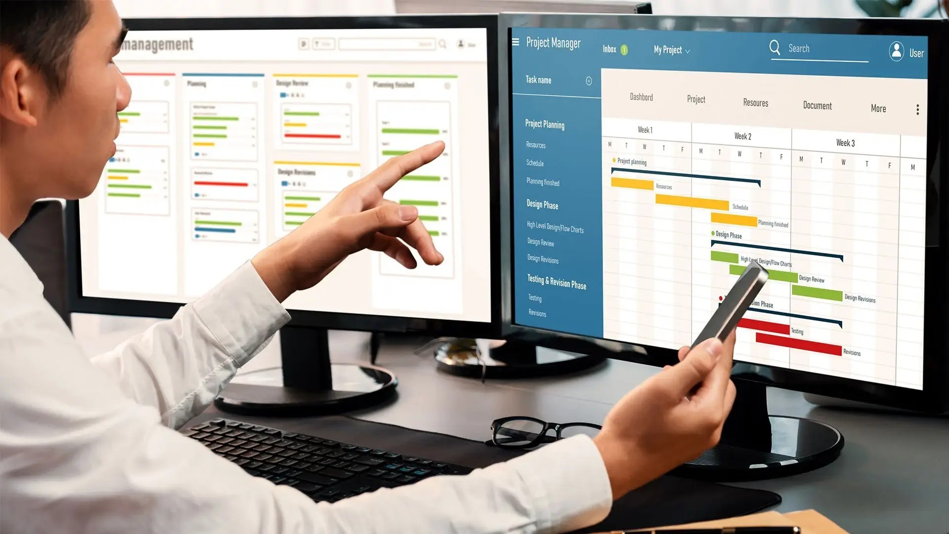 A person in a white shirt is seated at a desk, pointing at a computer screen displaying a project management dashboard with various colored bars representing scheduling and tasks. The individual is also holding a mobile phone in the other hand and there are a pair of glasses on the desk.