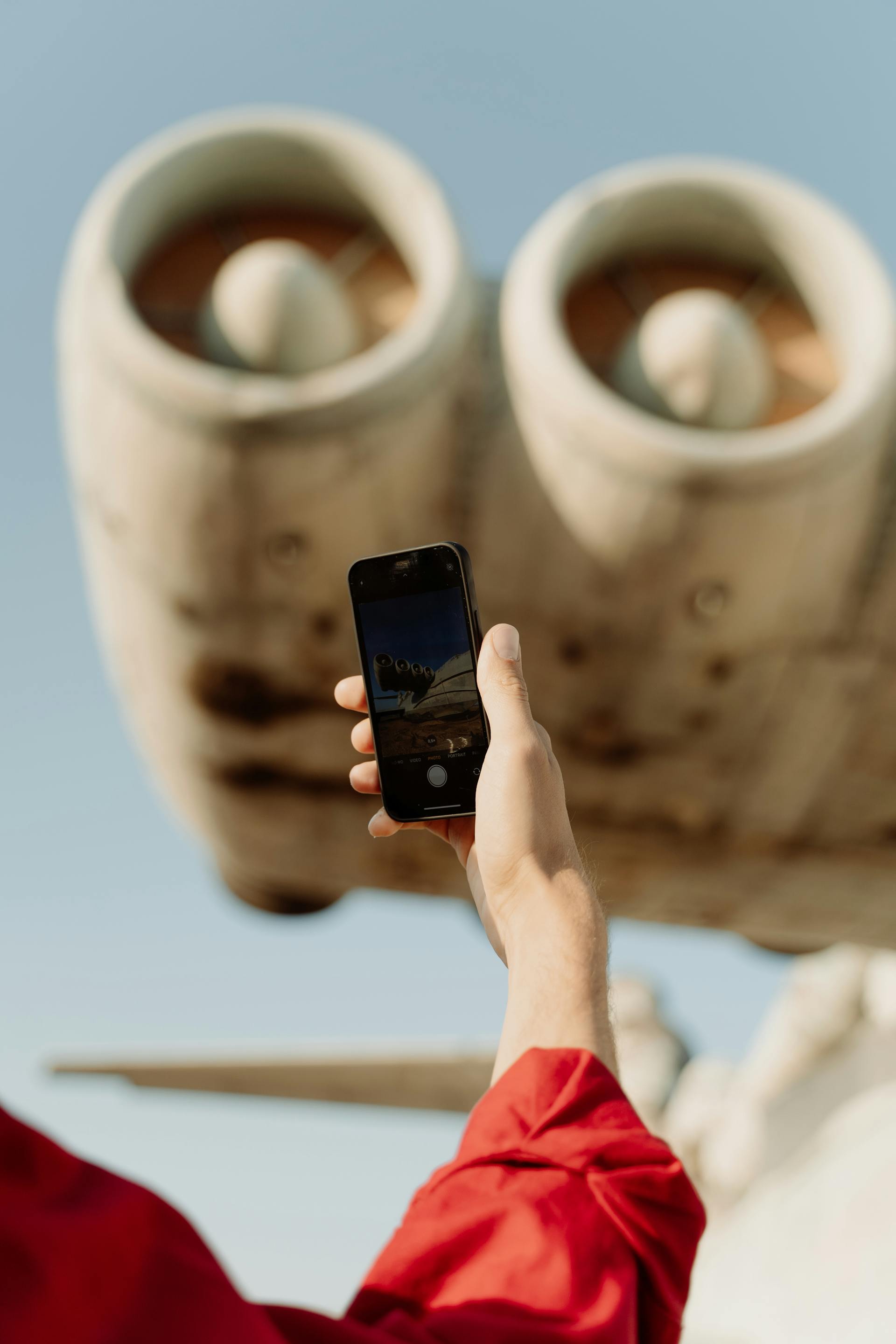 A person in a red shirt holds up a smartphone to take a photo of the large jet engines of an aircraft, with the camera screen showing the live view of the plane.