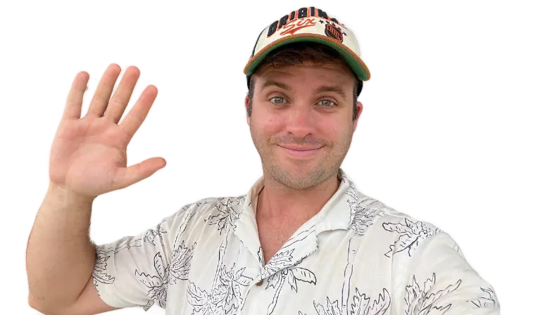 A smiling person in a hat waves with one hand, wearing a light-colored shirt against a neutral background.
