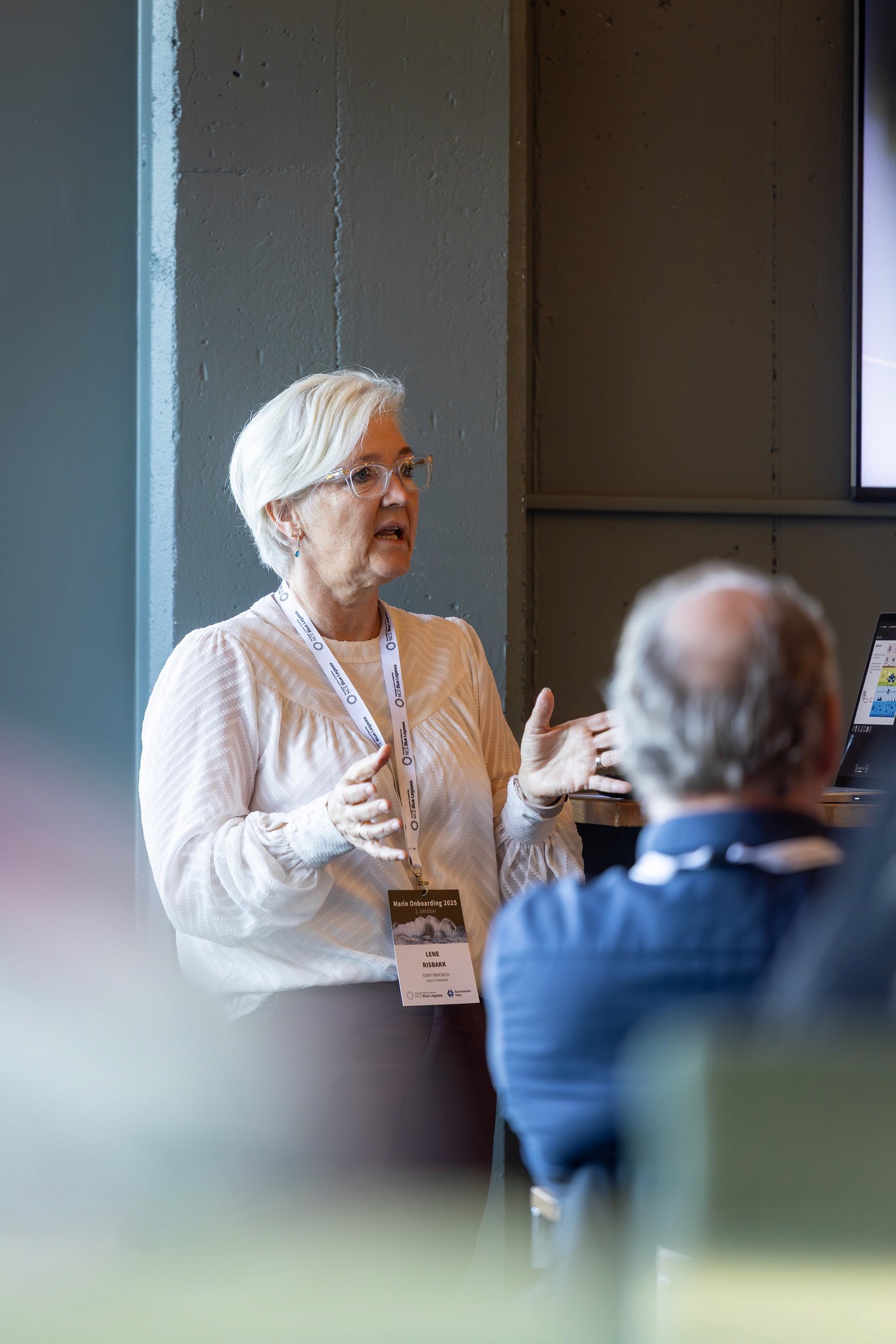 A person with light hair gestures while speaking to an audience in a modern setting.