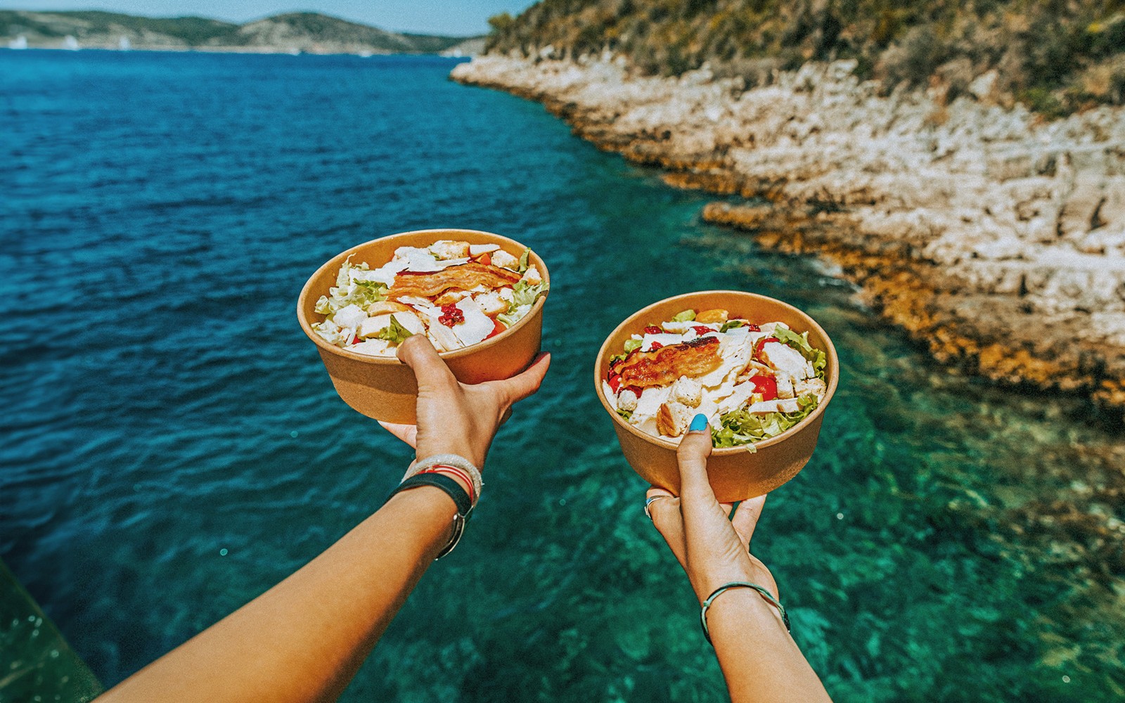 Hands holding salad bowls over clear blue water on a Trogir to Hvar boat cruise.