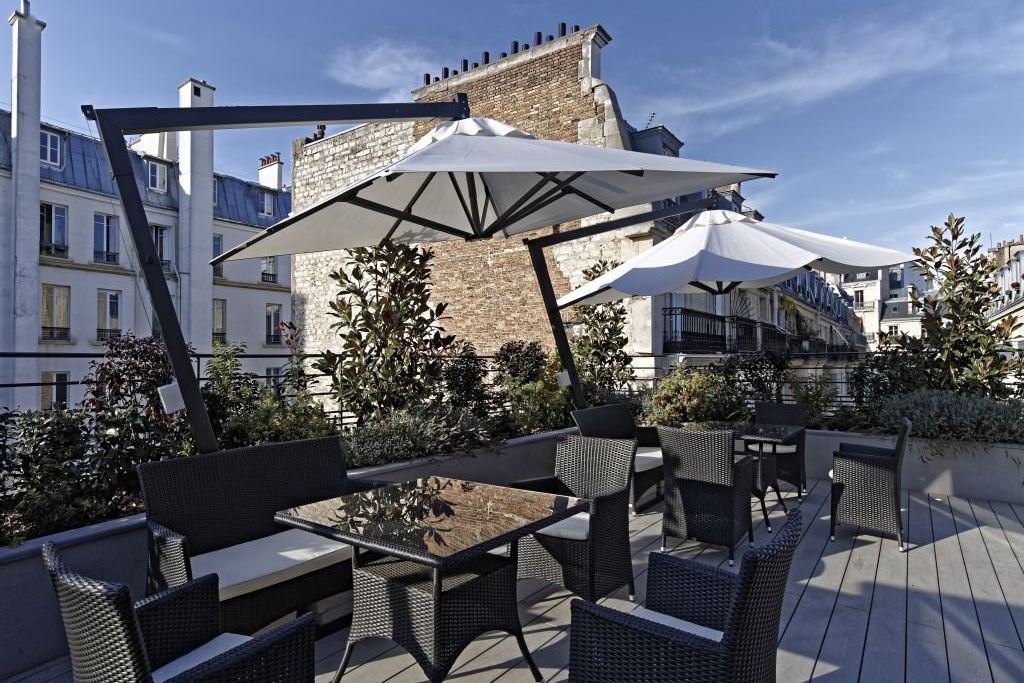 Spacious rooftop terrace with modern parasols, multiple seating areas in black wicker, raised planters, and surrounding Parisian architecture under blue sky.