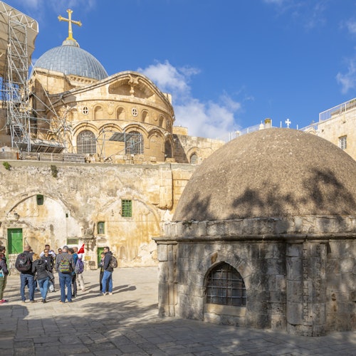 A group of tourists stand near the Church of the Holy Sepulchre in Jerusalem, with its domes and surrounding stone structures visible.