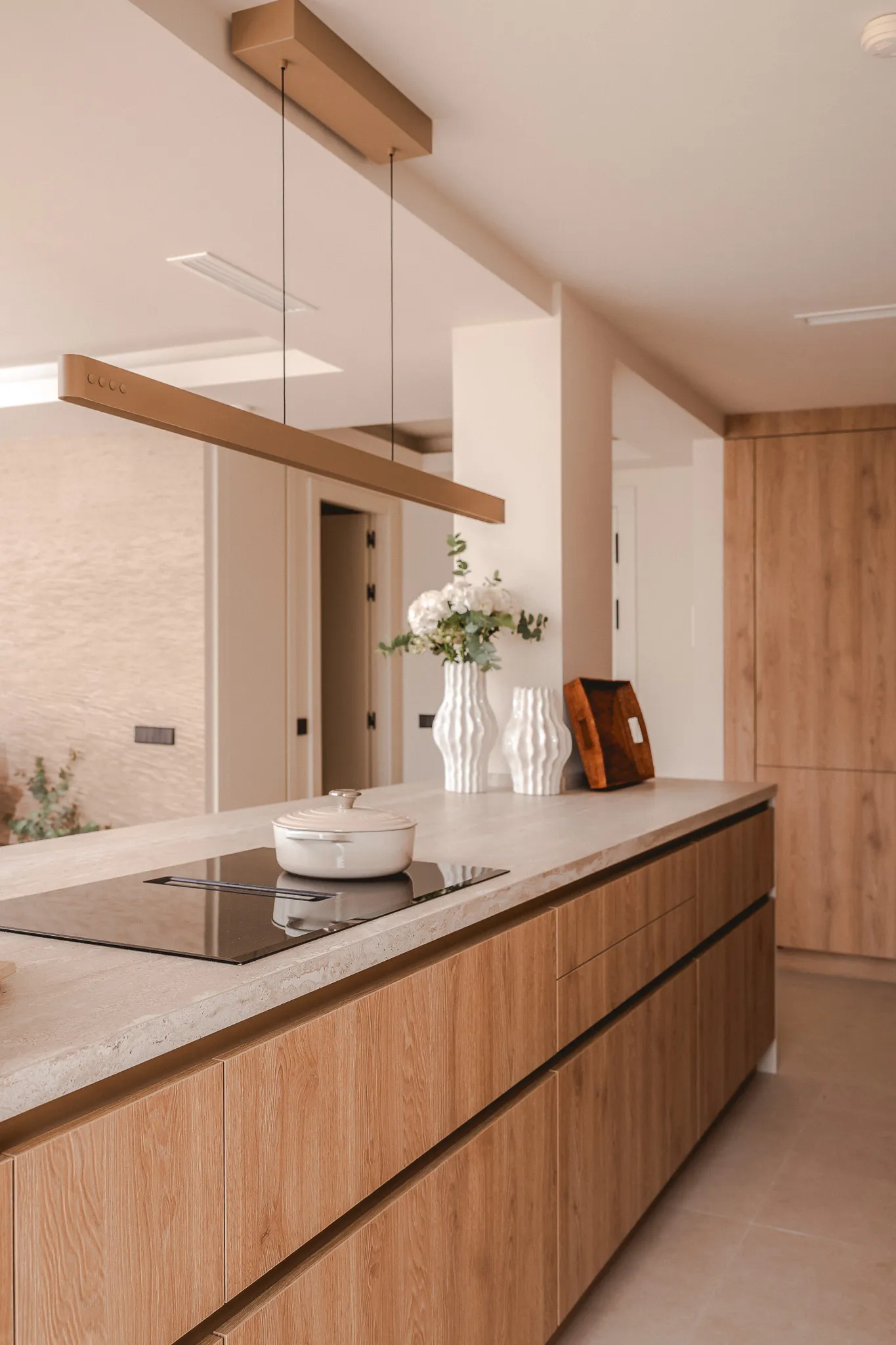 A close-up of a modern light wood kitchen island featuring an integrated cooktop with a white ceramic pot and a sleek linear pendant light hanging above.