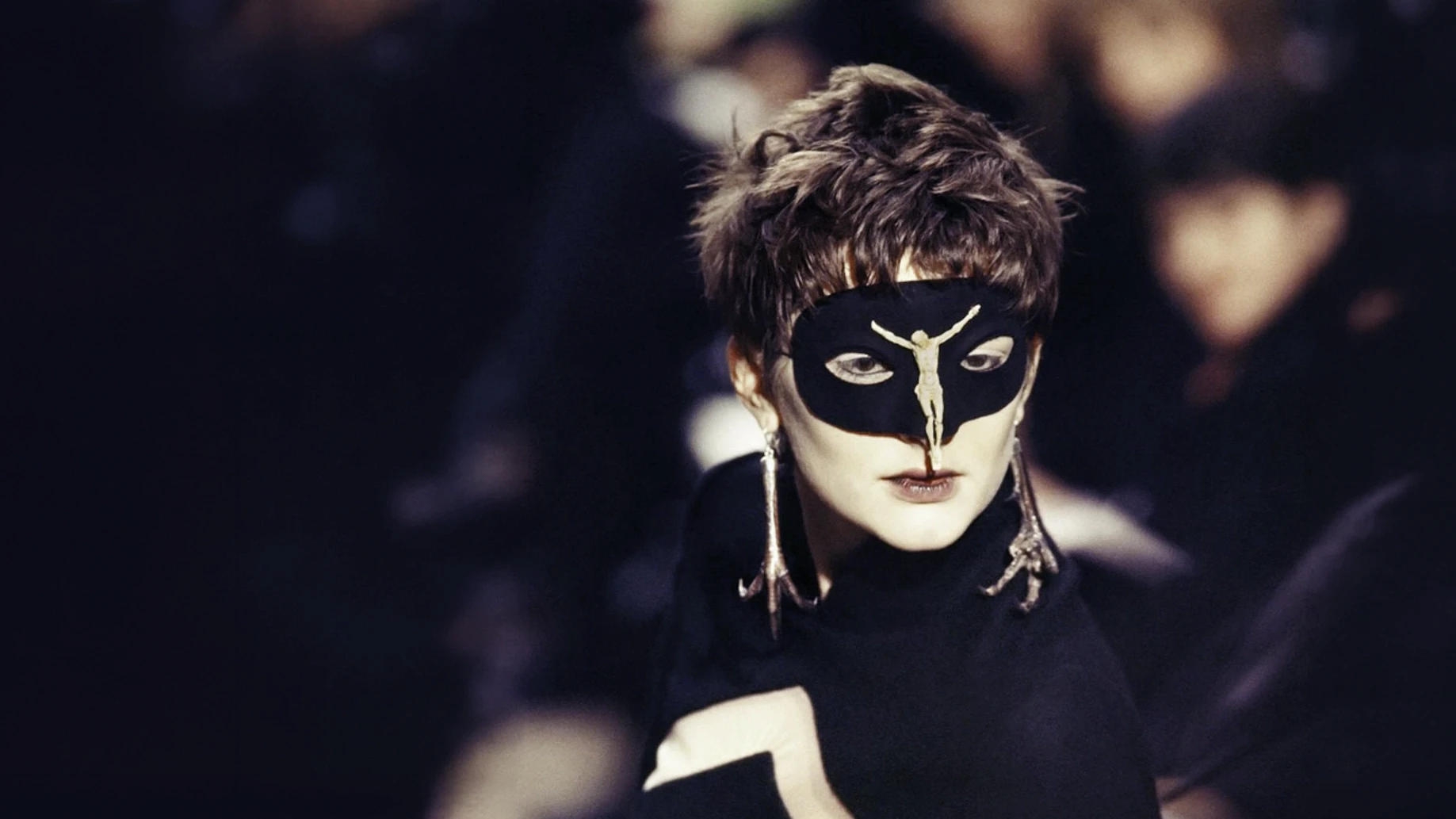 Model on a dark runway wearing a black lace mask and a silver pheasant claw earring designed by Shaun Leane for the Alexander McQueen Dante collection.