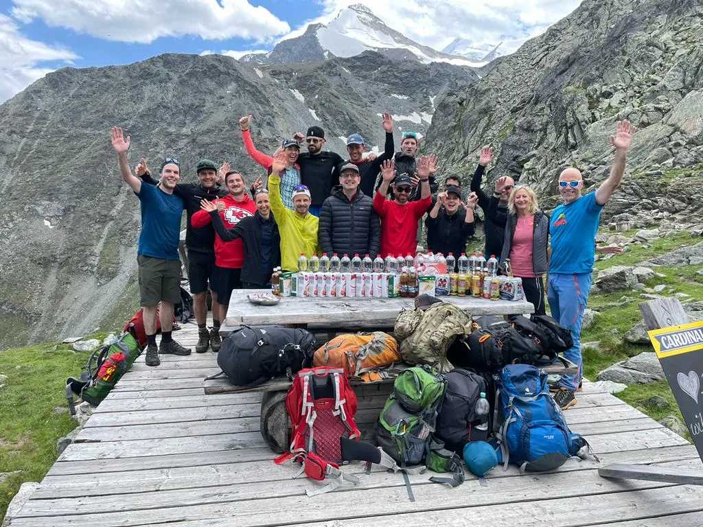 Wandergruppe mit Rucksäcken und Getränken auf einer Holzplattform vor Gletscher und Berggipfeln