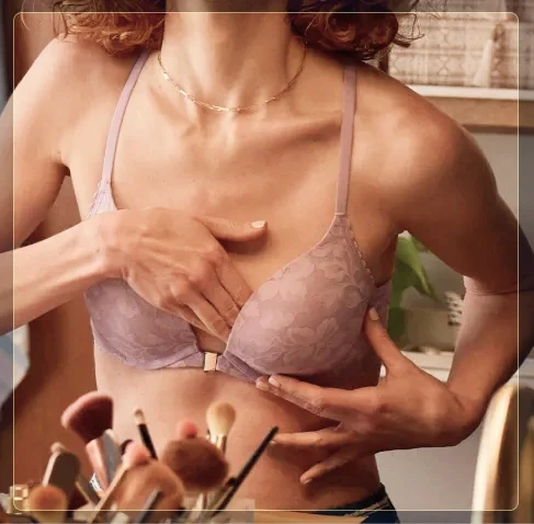 A woman in a lavender lace bra performs a breast self-exam in the mirror in a softly lit room.