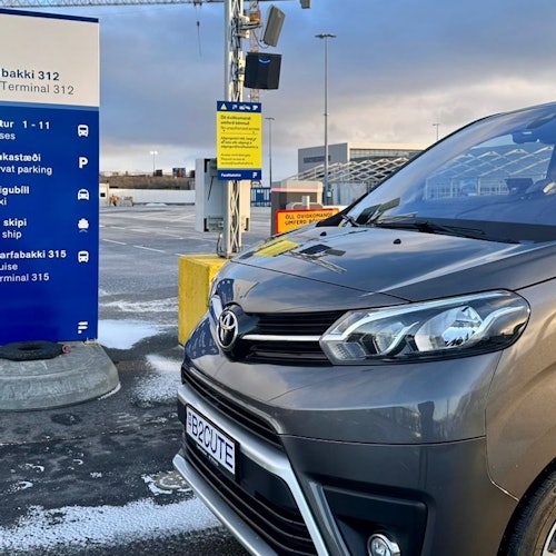 Una furgoneta Toyota gris con la matrícula "B8CUTE" estacionada junto a un letrero azul de información en una terminal del aeropuerto bajo un cielo nublado.