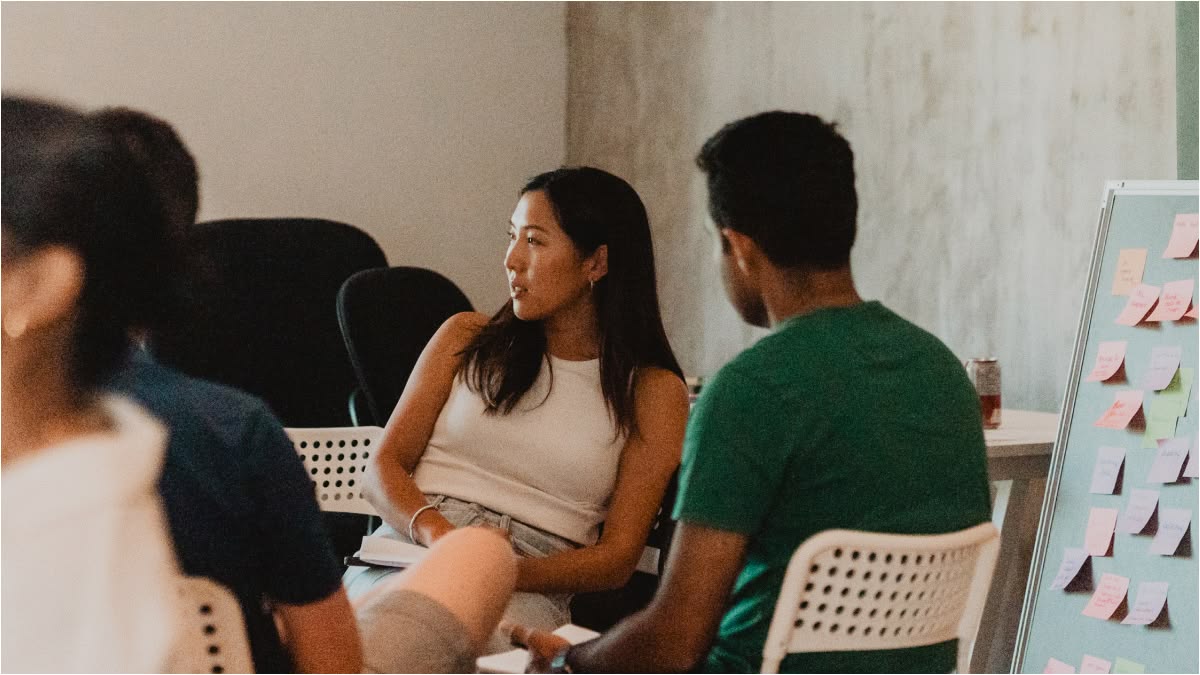 A group of people sit in a casual office setting, engaged in discussion. Colorful sticky notes cover a board, creating a collaborative and focused atmosphere.
