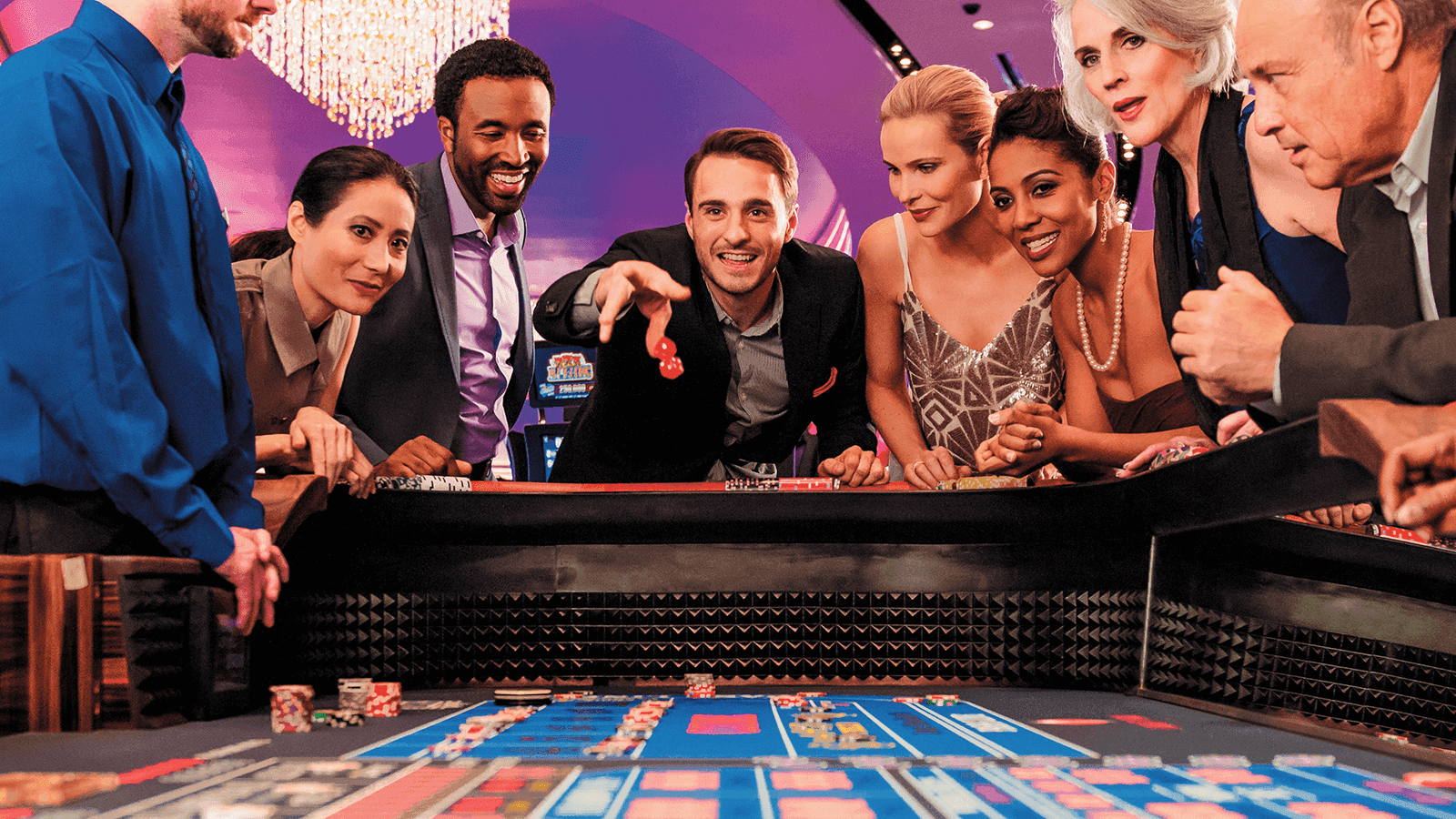 A group of people gather around a casino craps table while a man in the center throws dice, with chips and cards visible on the table.