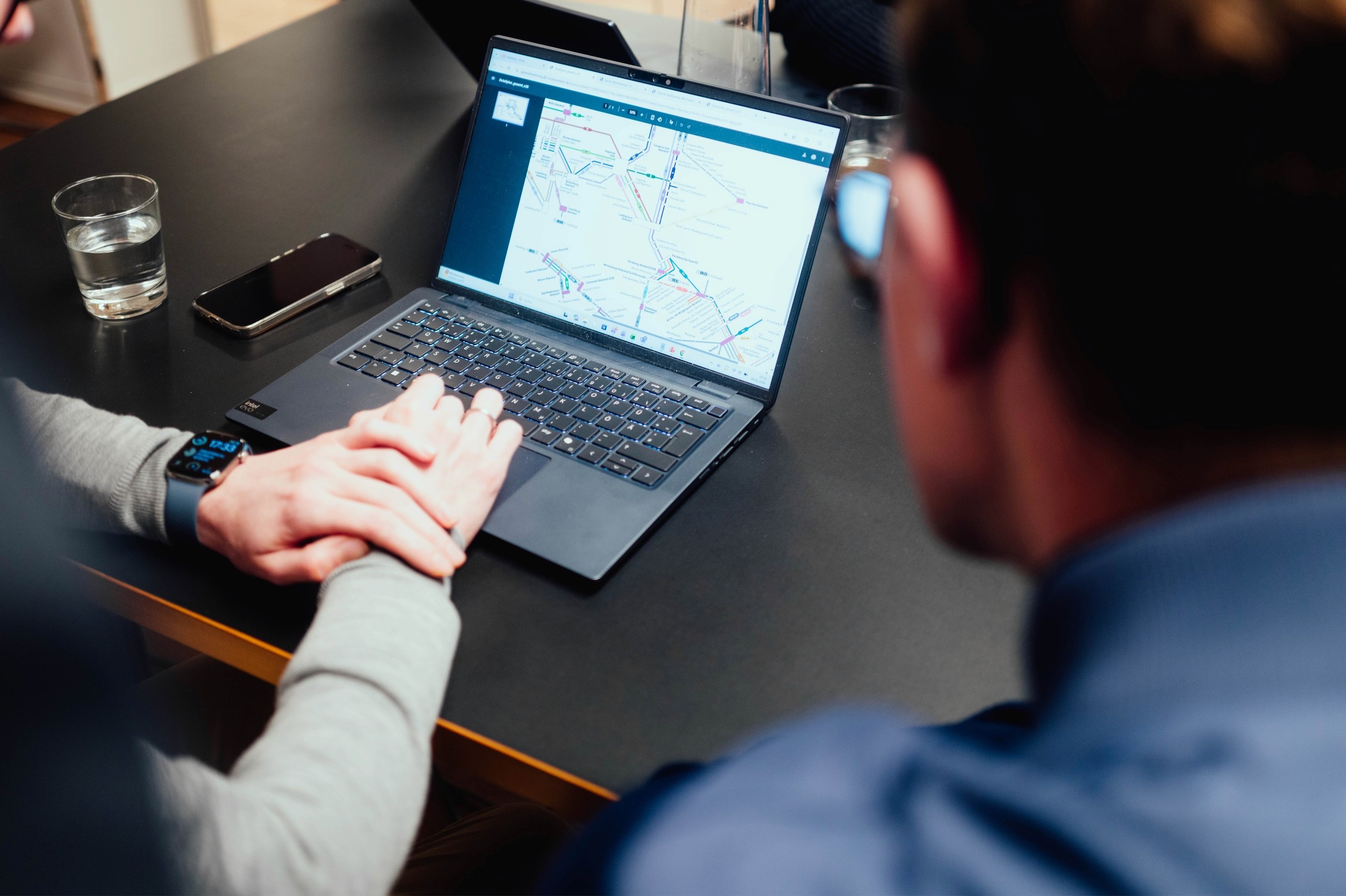 - Zwei Personen begutachten eine Karte oder ein Diagramm auf einem Laptop, mit einem Wasserglas und Smartphone auf einem dunklen Tisch; dies steht für konzentrierte Diskussion und strategische Planung während einer Beratung.
