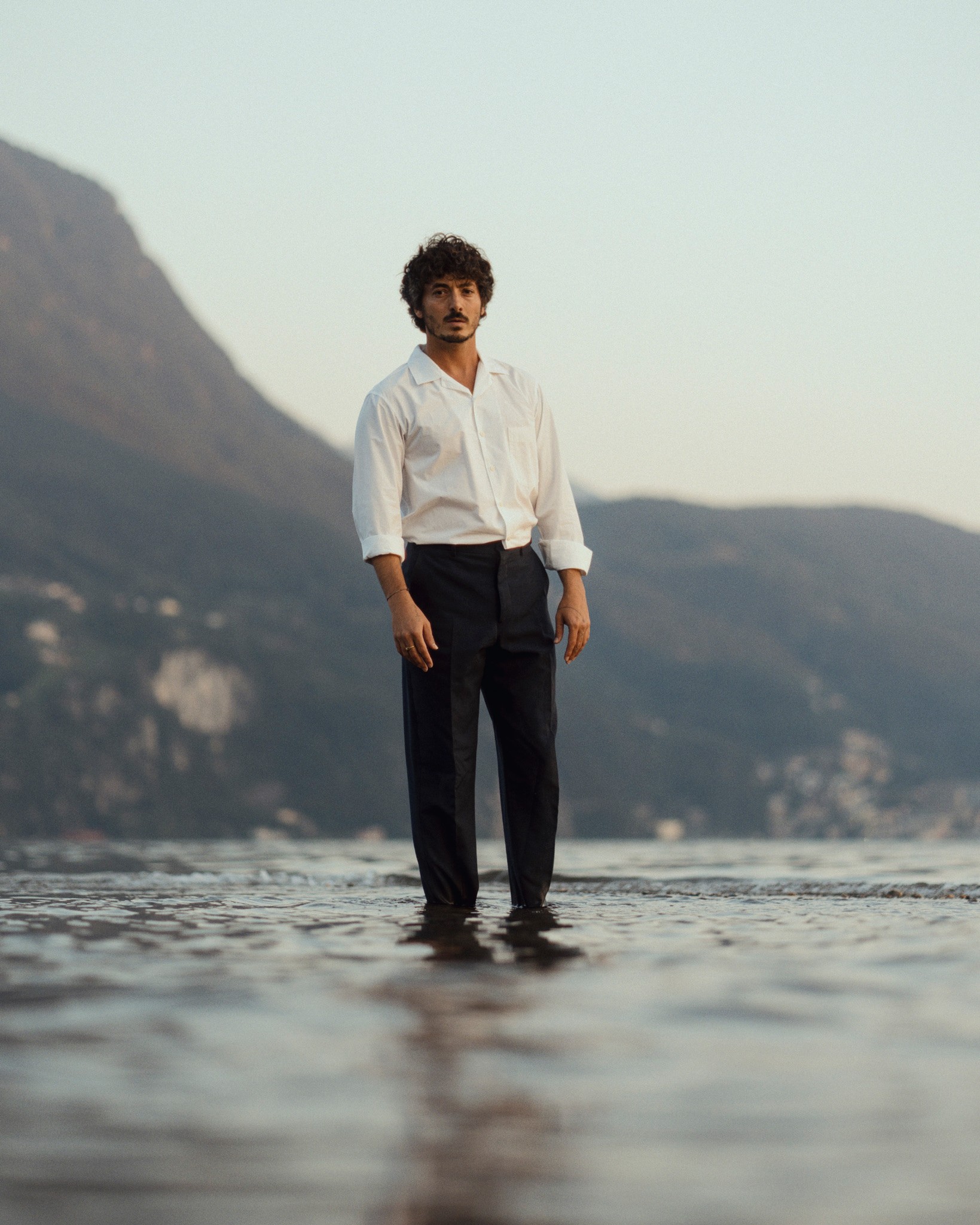 Portrait de l'artiste italien Lumi habillé en chemise blanche et pantalon de cosutme fixant l'ojectif et immobile sur l'eau lui donnant l'impression de marcher sur le lac de Lugano