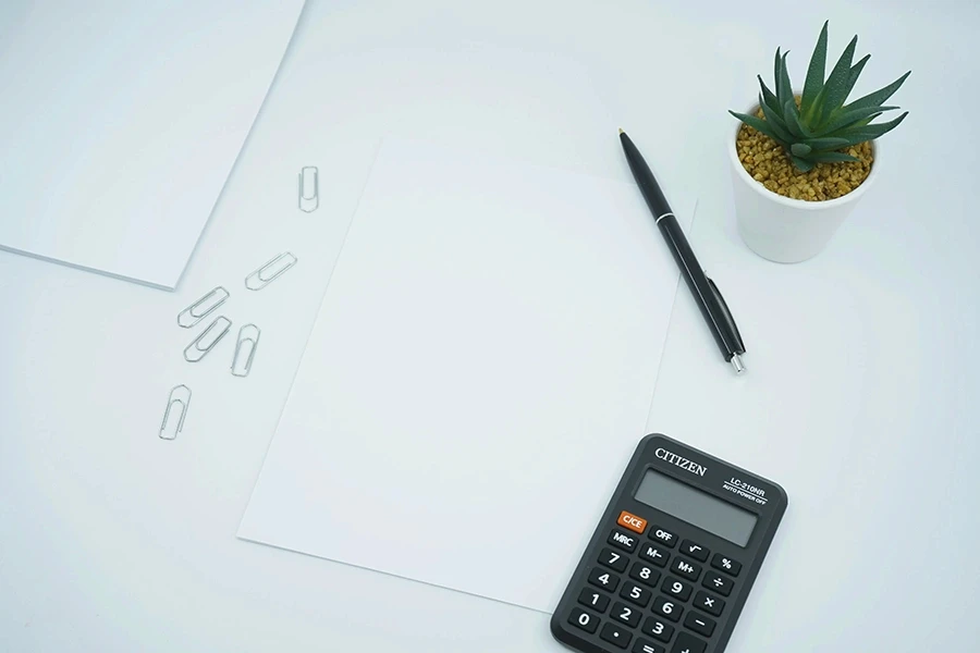 Minimalist workspace with blank sheets of paper, a black calculator, a black pen, scattered paper clips, and a small potted succulent plant on a white desk.
