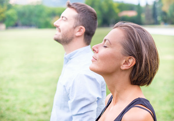 Two adults standing outdoors with eyes closed, facing upward, breathing fresh air in a green park setting.