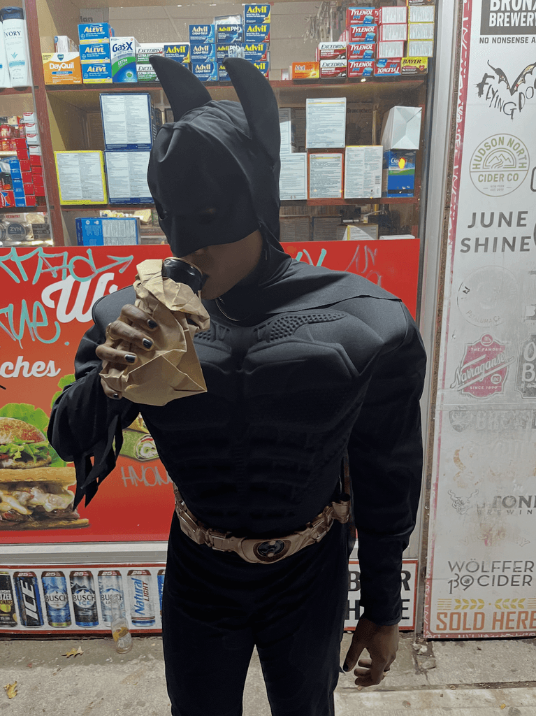 Ashleigh Warren in a Batman costume is holding a sandwich in front of a shop with colorful advertisements.