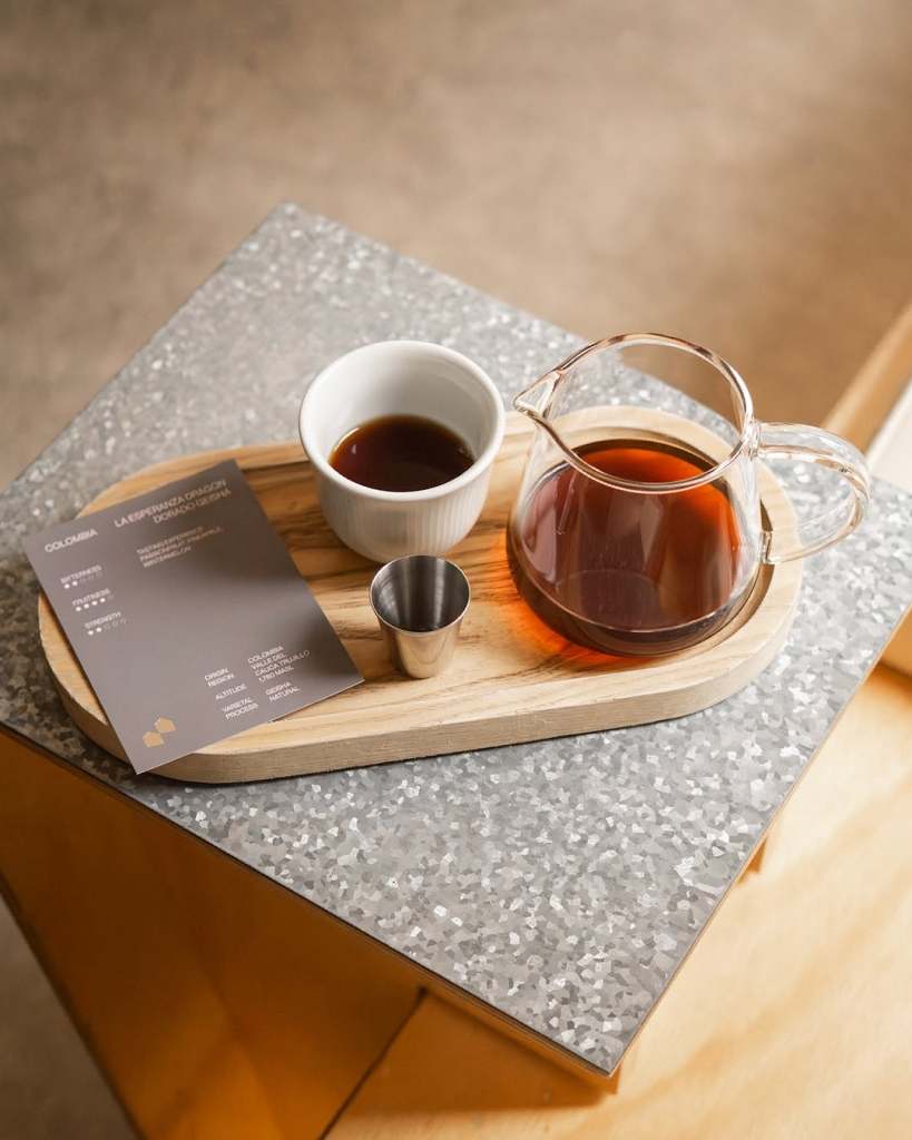 A wooden tray featuring a glass coffee carafe, a white cup of coffee, a metal shot glass, and a menu card on a textured grey surface.