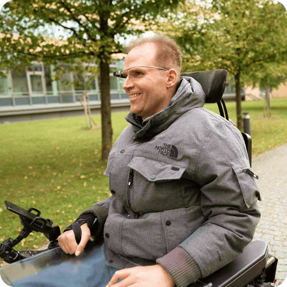 The image shows a man in a motorized wheelchair, wearing munevo smart glasses, which he uses to control the movement of his wheelchair. He appears outdoors, dressed in a grey jacket, with a cheerful expression. The background features green trees and a building, suggesting an outdoor or campus environment. The use of these smart glasses emphasizes their role in enhancing mobility and independence for individuals with mobility challenges.