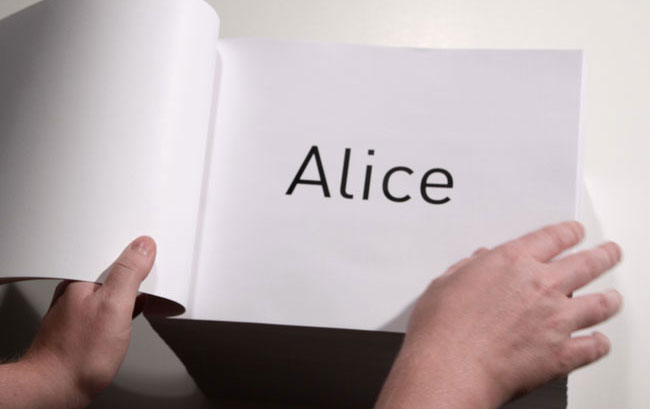 Two hands are shown holding open a white book with the word "Alice" printed in black on the page. The book is open toward the viewer, and the hands are positioned on either side of the open pages. The background is a plain white wall.