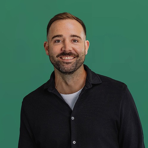 Smiling man with short hair and a mustache wearing a black button-up shirt against a green background.