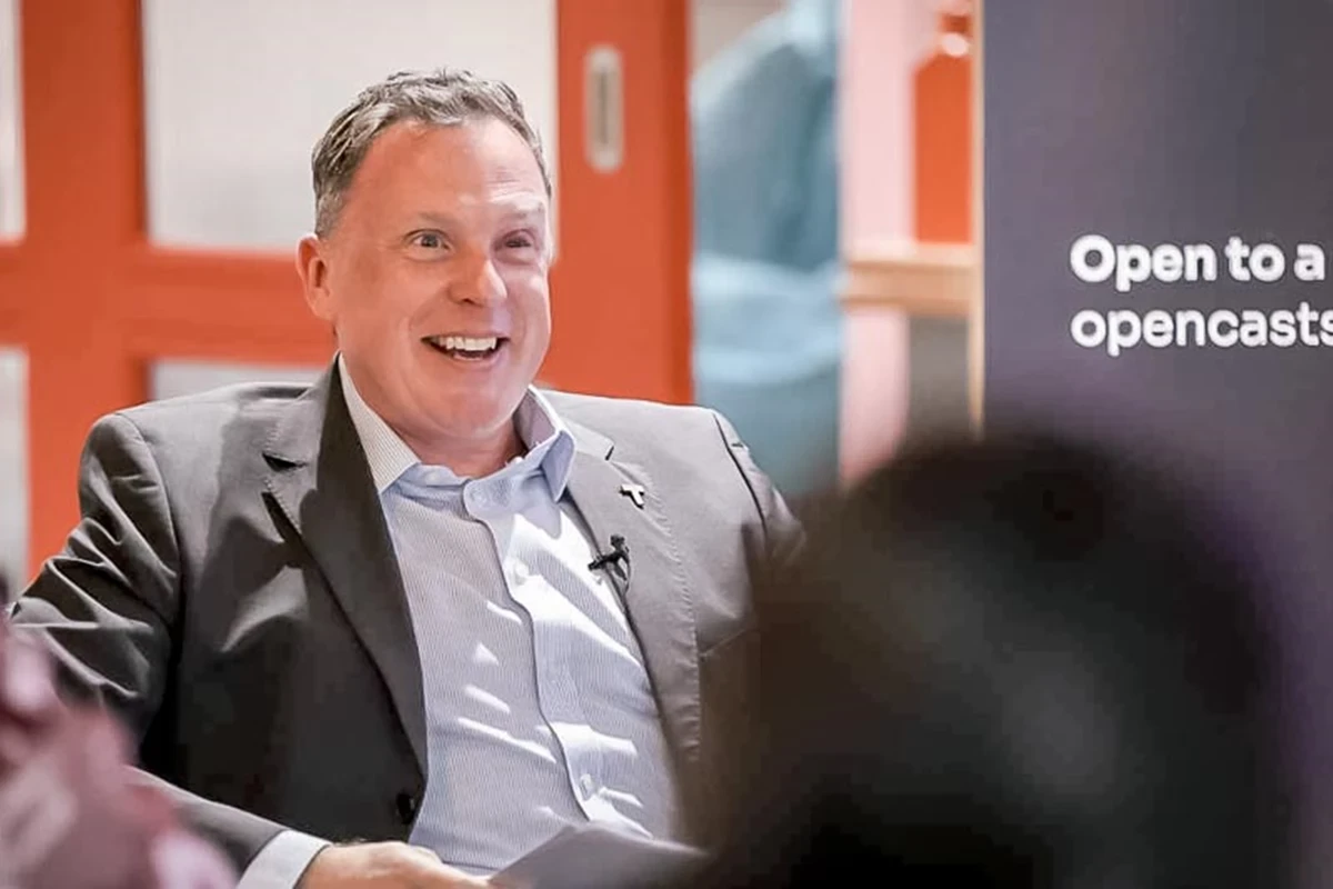 Three panel speakers sit on an orange sofa behind a low table with books, water glasses, and a sign displaying a QR code. The setting suggests a moderated discussion or talk at a public event.