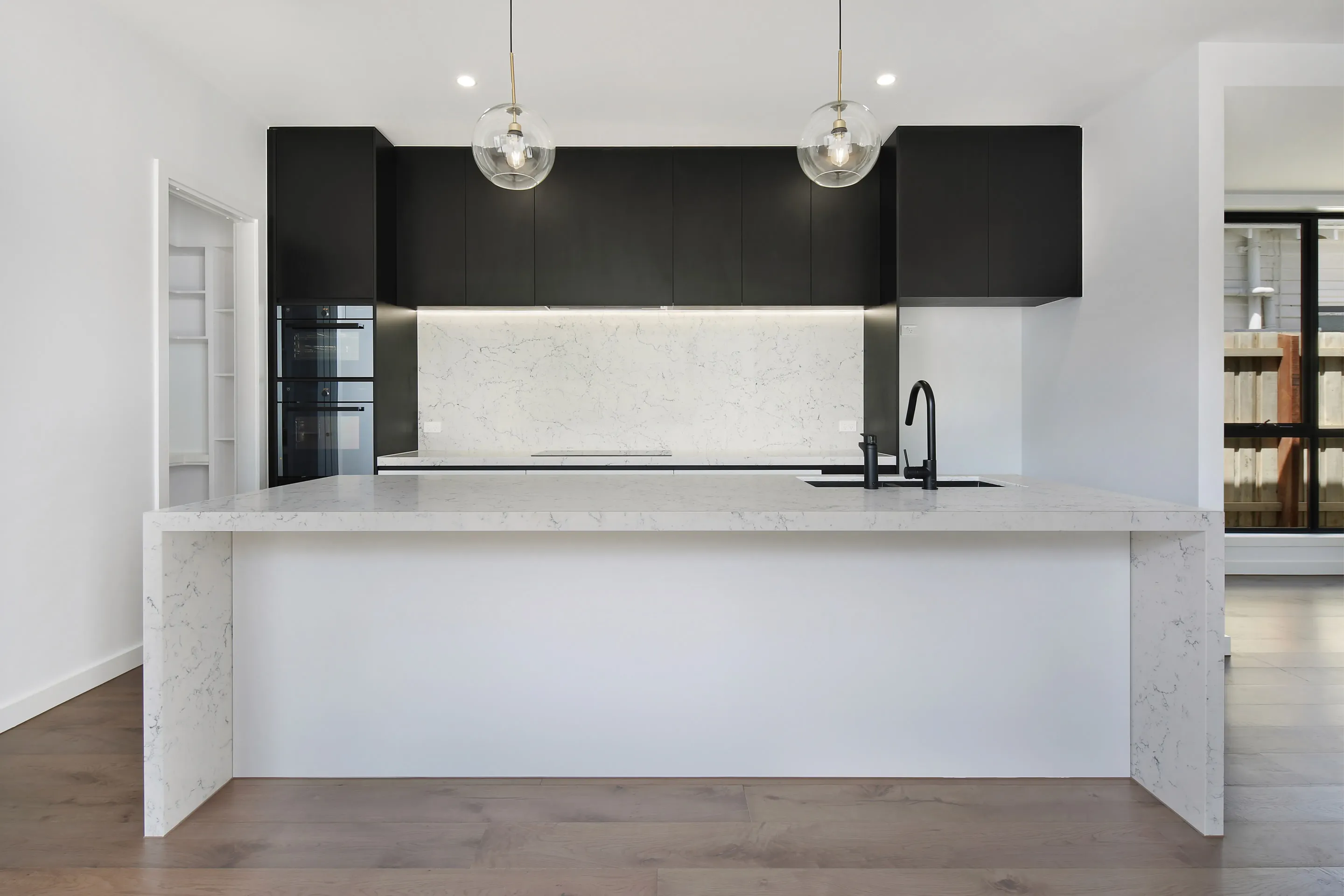 Kitchen with matte-black overhead cabinetry, two clear-glass globe pendants, a veined white stone splashback and waterfall island, black tap and oak floors