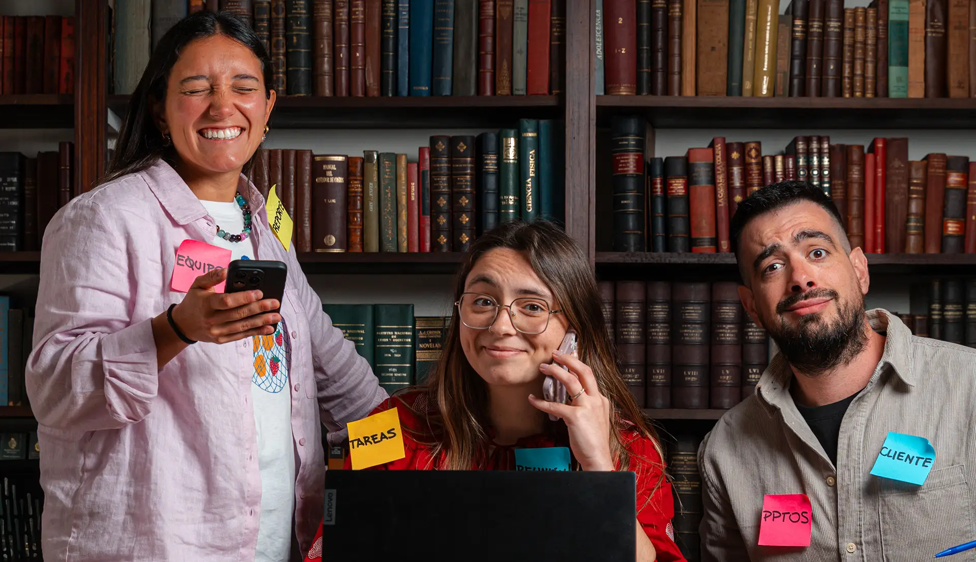 Retrato frontal de dos chicas y un chico, trabajando en conjunto en una computadora, con post its pegadas sobre ellos. Una de las chicas habla por teléfono y la otra mira la pantalla de su celular. Tienen expresiones graciosas en sus caras. De fondo se ve una biblioteca con un montón de libros.