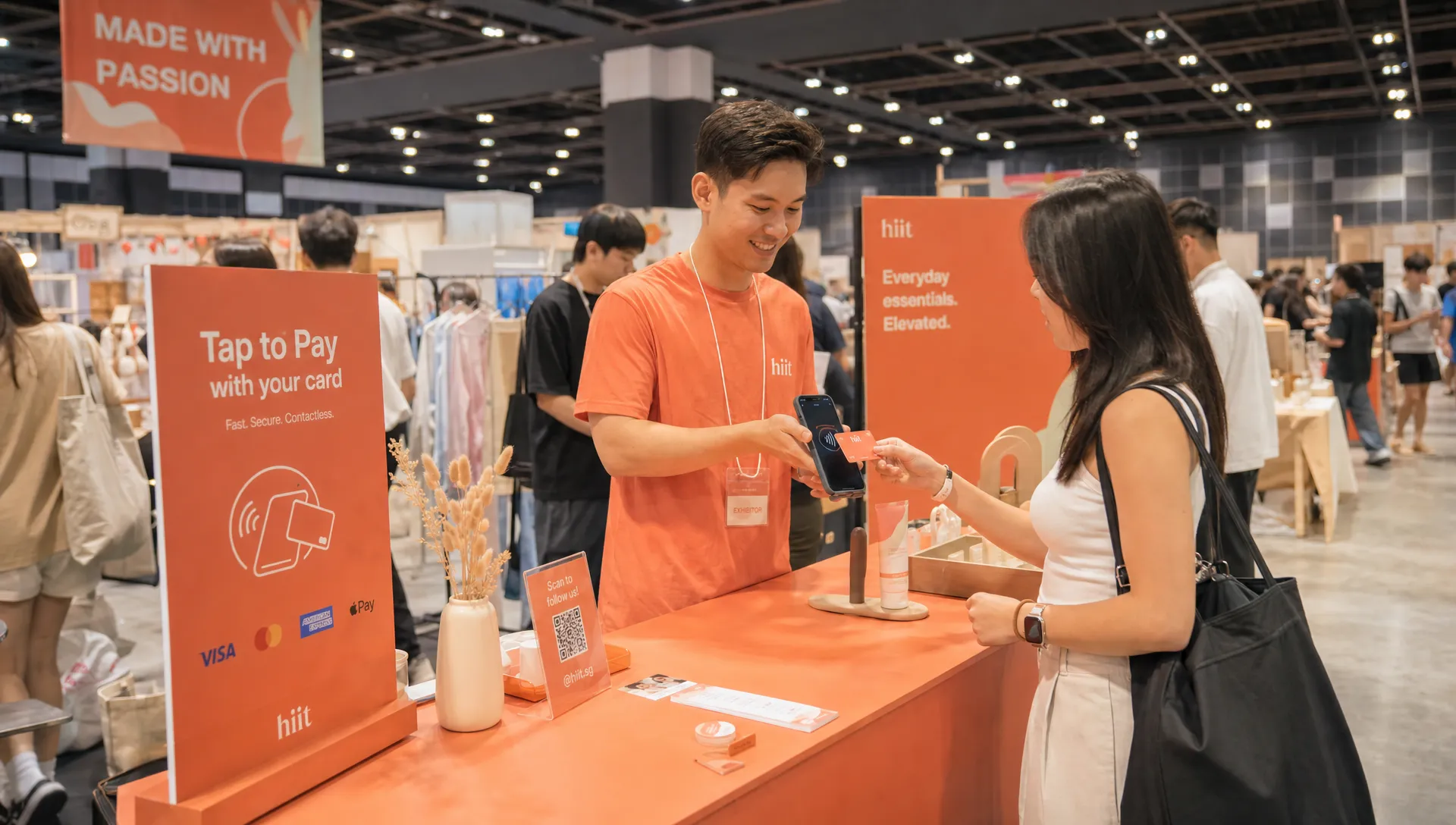 A Singapore pop-up booth where a customer taps a contactless card on the back of a merchant's smartphone, with product displays on the table and no separate card terminal visible, seen from a slightly wider angle with more of the event hall around the checkout area.