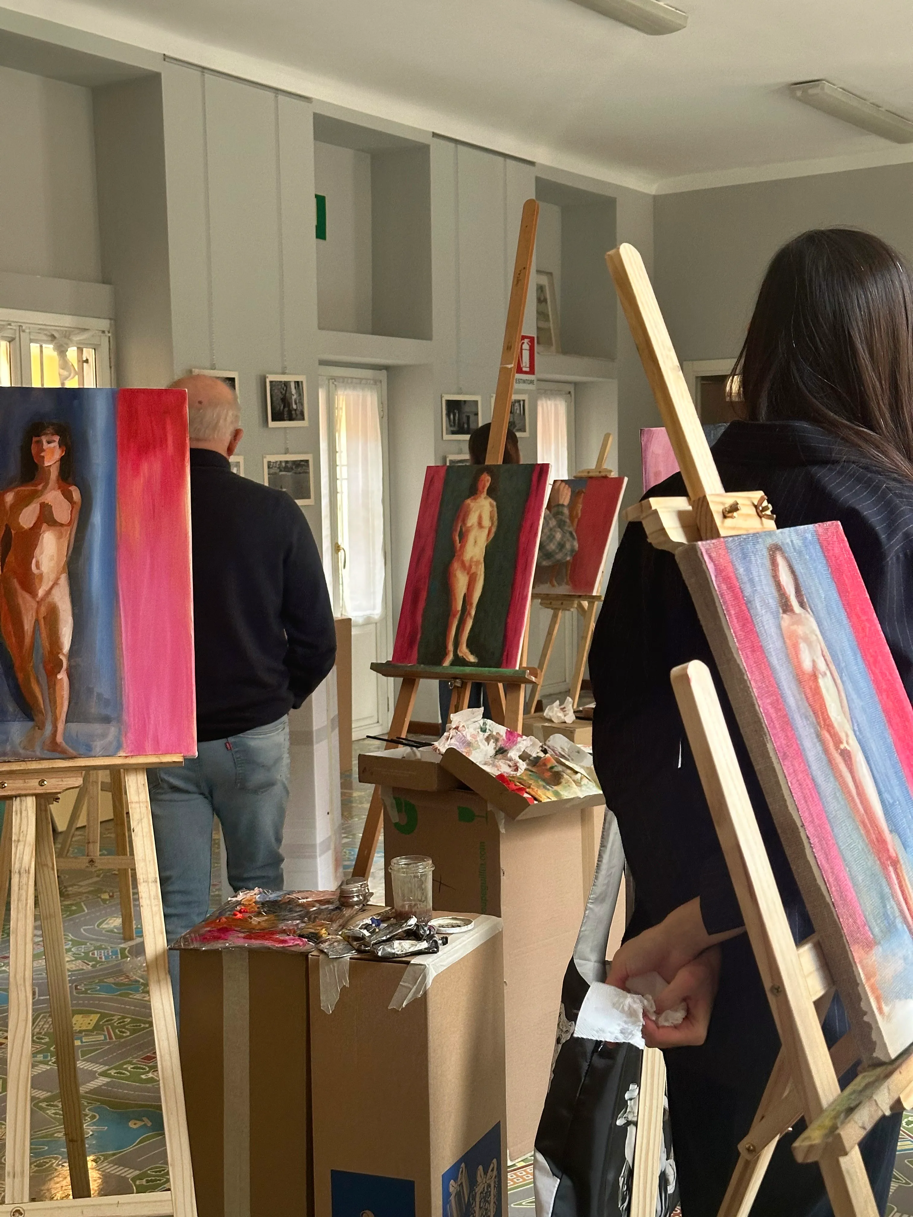 In a well-lit art studio, two artists focus intently on painting a male model posed against a multicolored draped backdrop, emphasizing the creative process and live figure drawing.