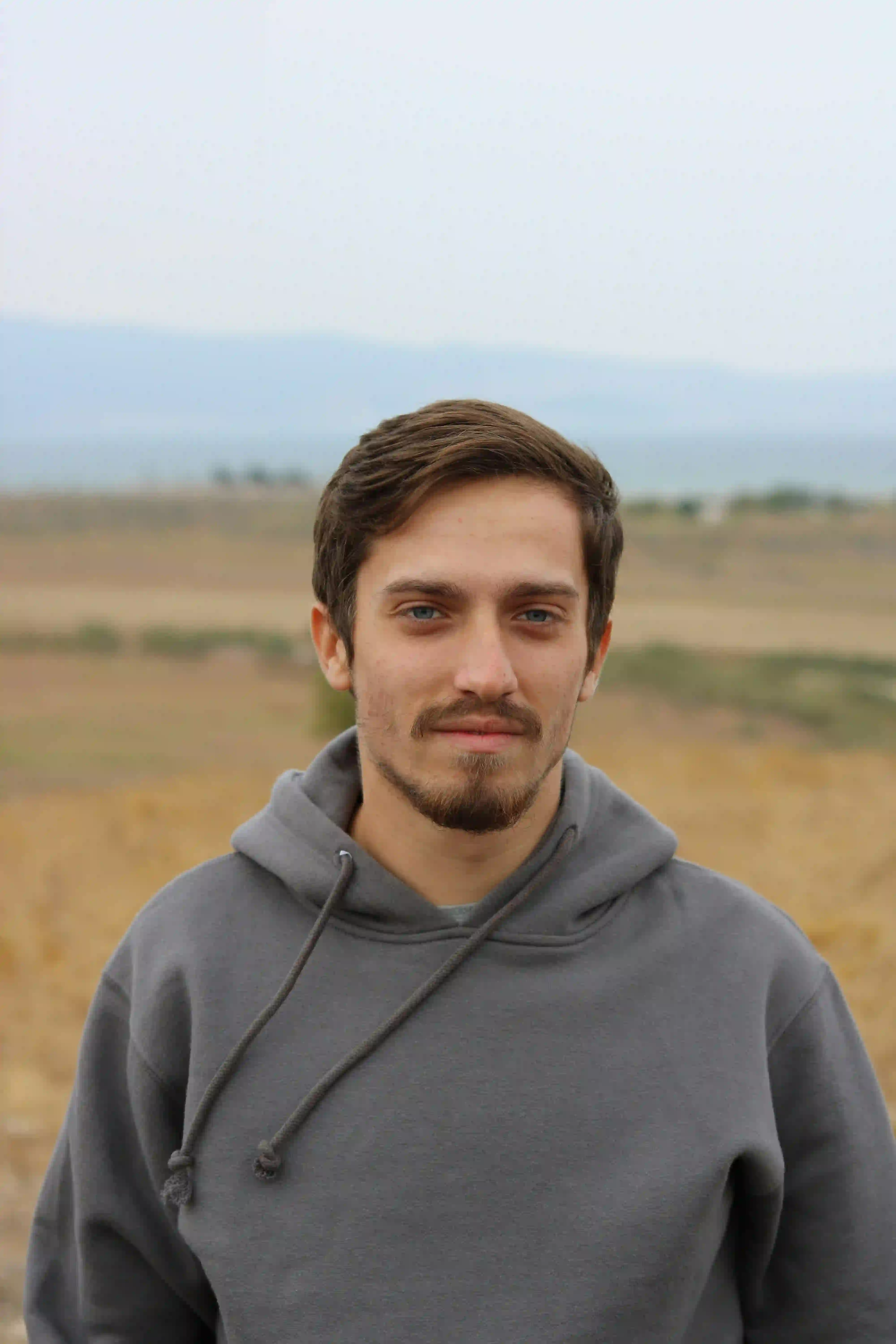 Young man in a gray hoodie standing outdoors with a blurred rural landscape in the background.