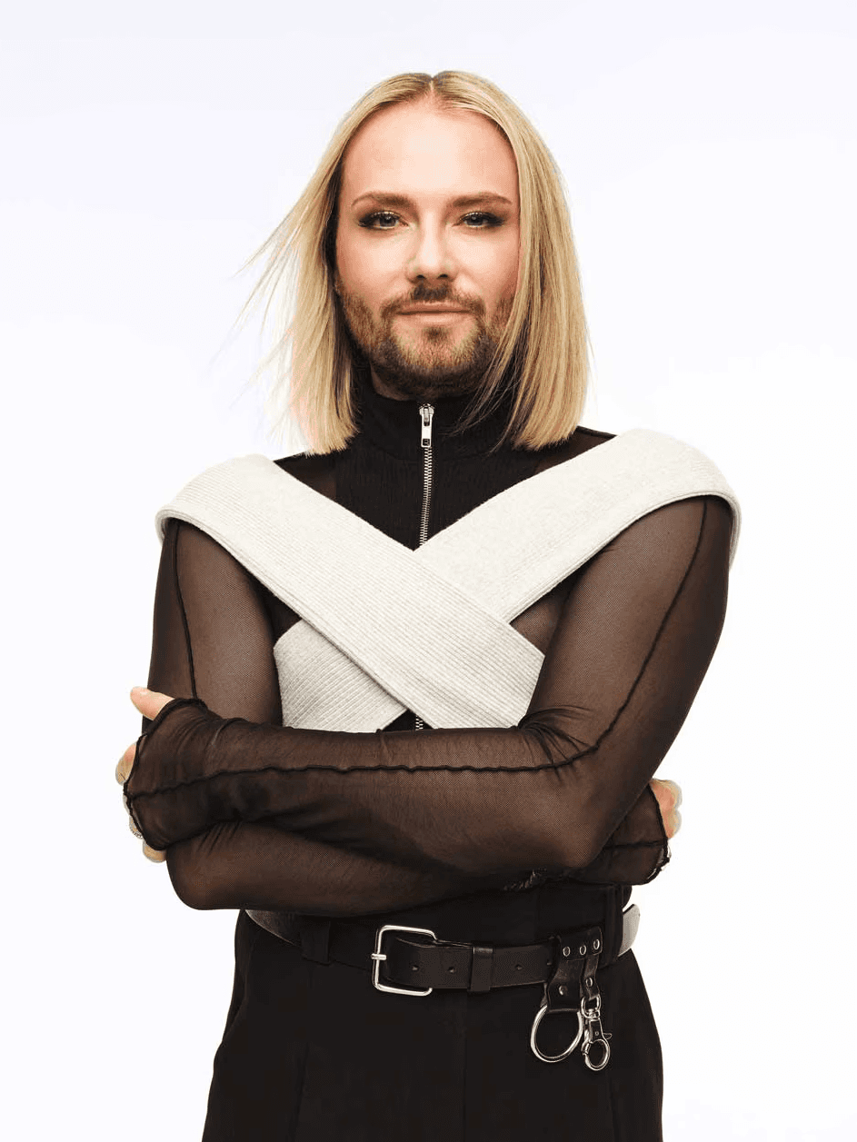 A person with shoulder-length blonde hair wears a stylish black and white outfit with crossed arms, set against a plain background.