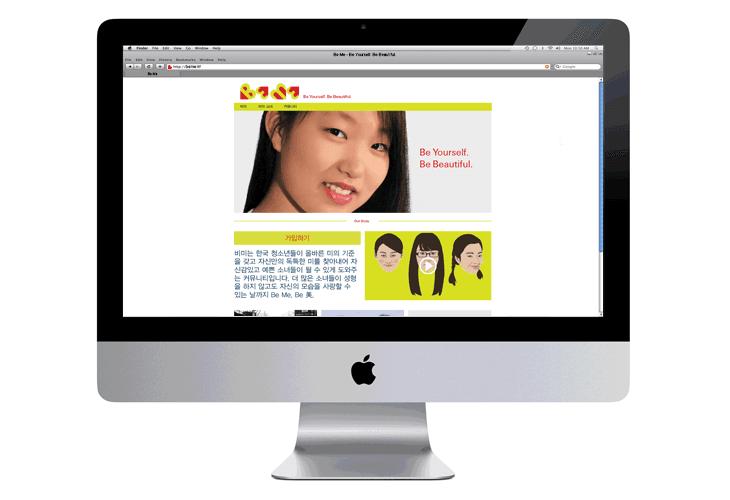 A silver Apple iMac computer displays a webpage with a Korean-language advertisement. The ad features a woman with long dark hair and a bright smile, alongside three smaller images of women with varying hairstyles and expressions. The webpage’s header includes “Be Yourself. Be Beautiful.” and a Google search bar. The computer’s screen is centered on the display.
