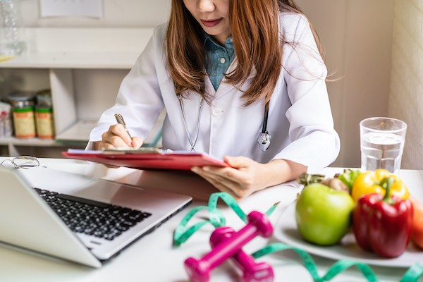 A person writing on a tablet beside a laptop, surrounded by fresh fruits and vegetables on a table.