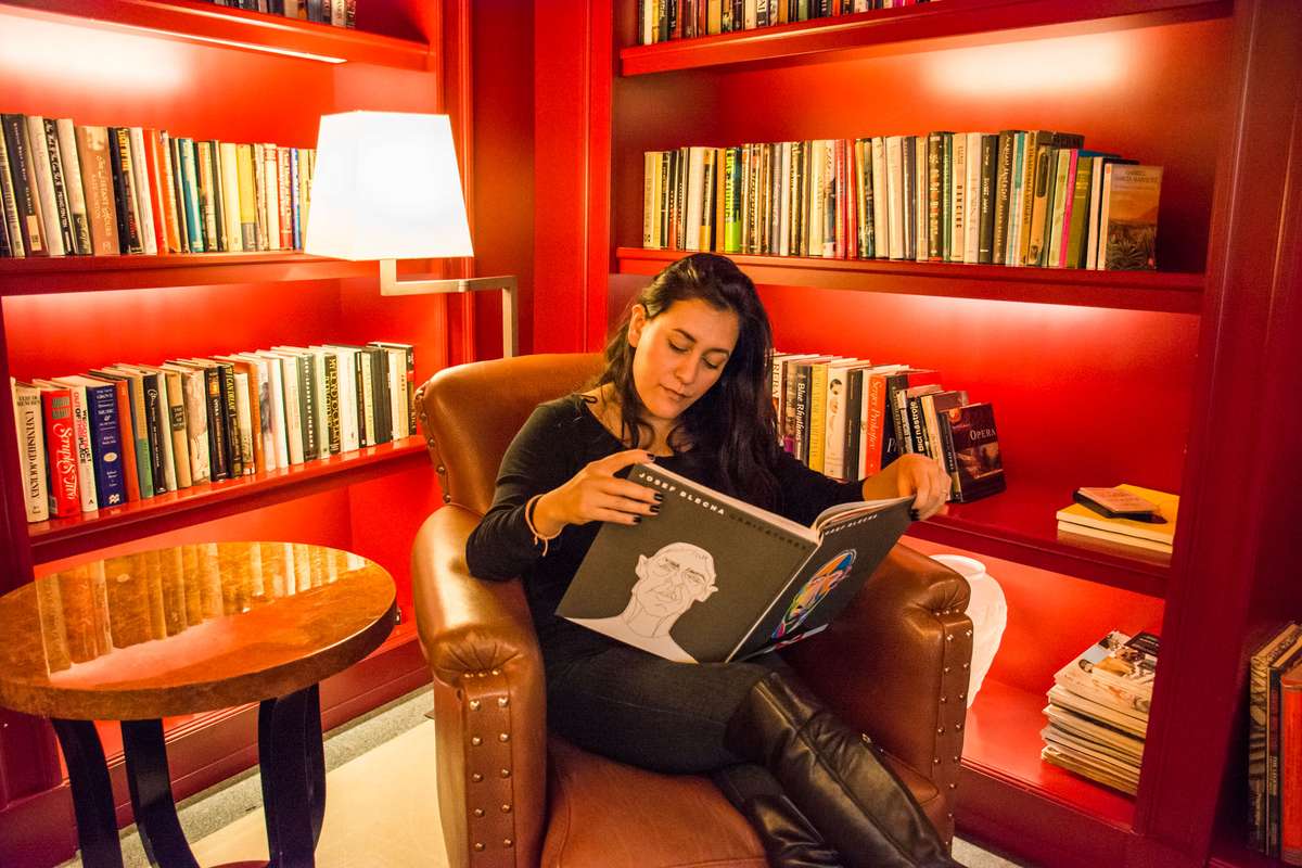 A woman sits in a leather armchair in a red library, reading a large book under a lamp surrounded by bookshelves.