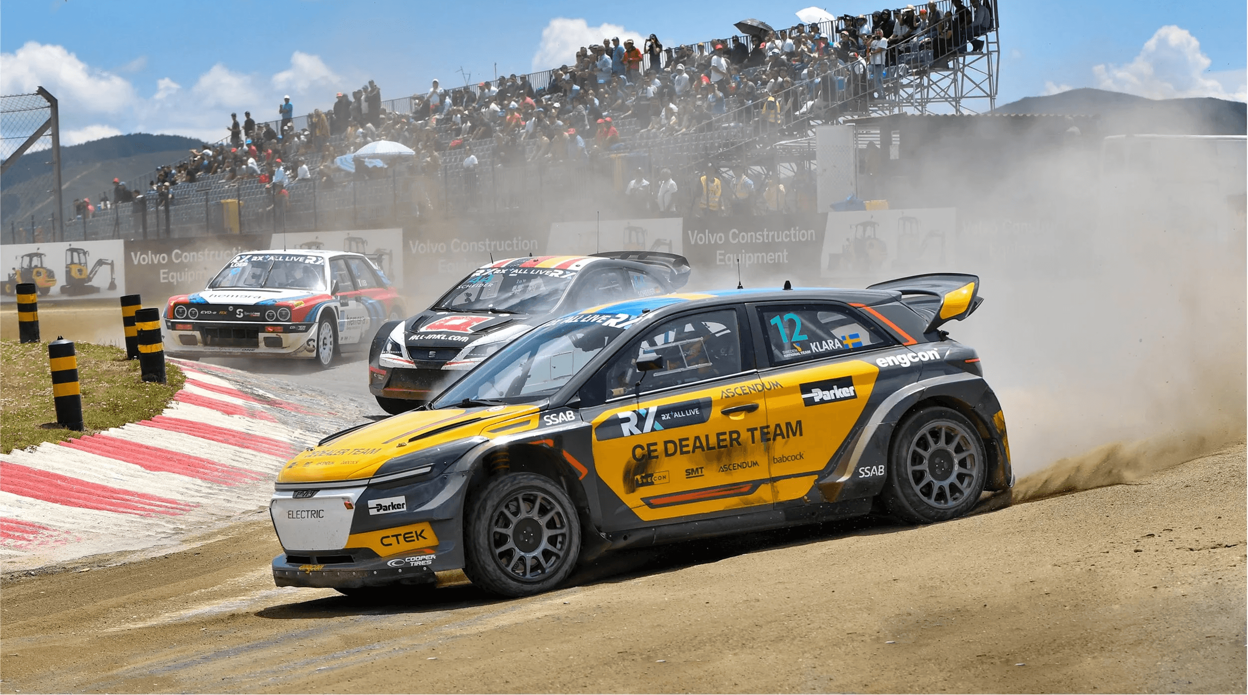 Three race cars drifting in a corner of a sandy track in front of a stand filled with the audience