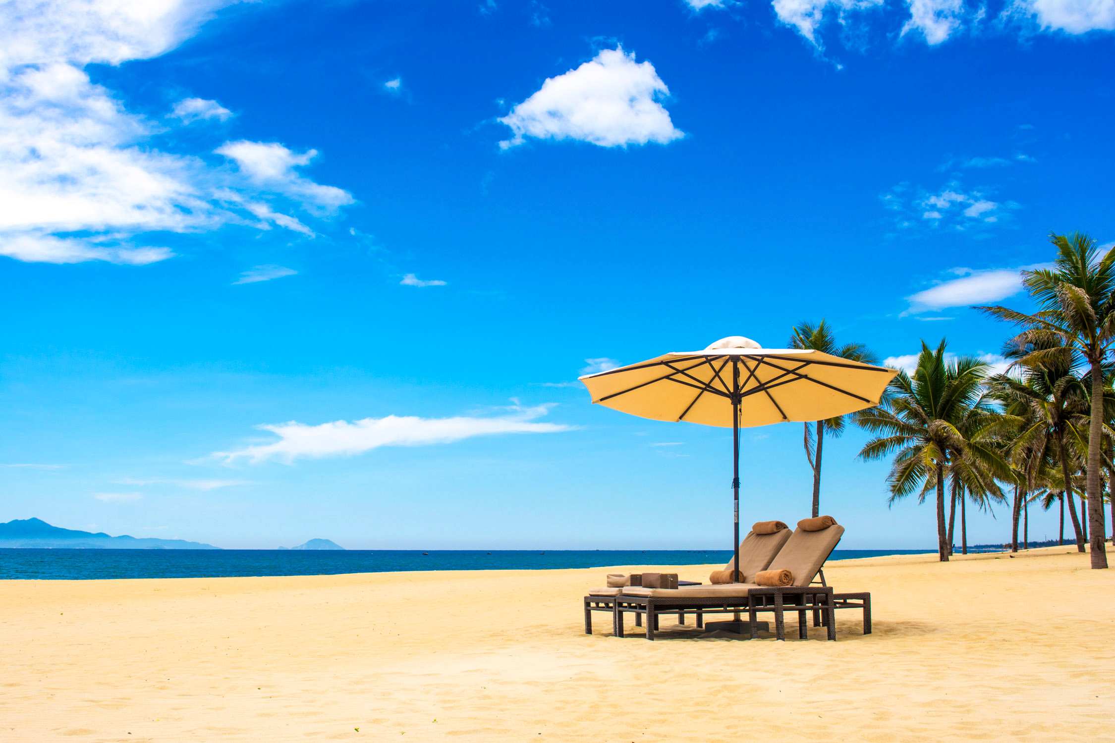 Two lounge chairs and a yellow beach umbrella sit on a sandy beach under a blue sky with white clouds.
