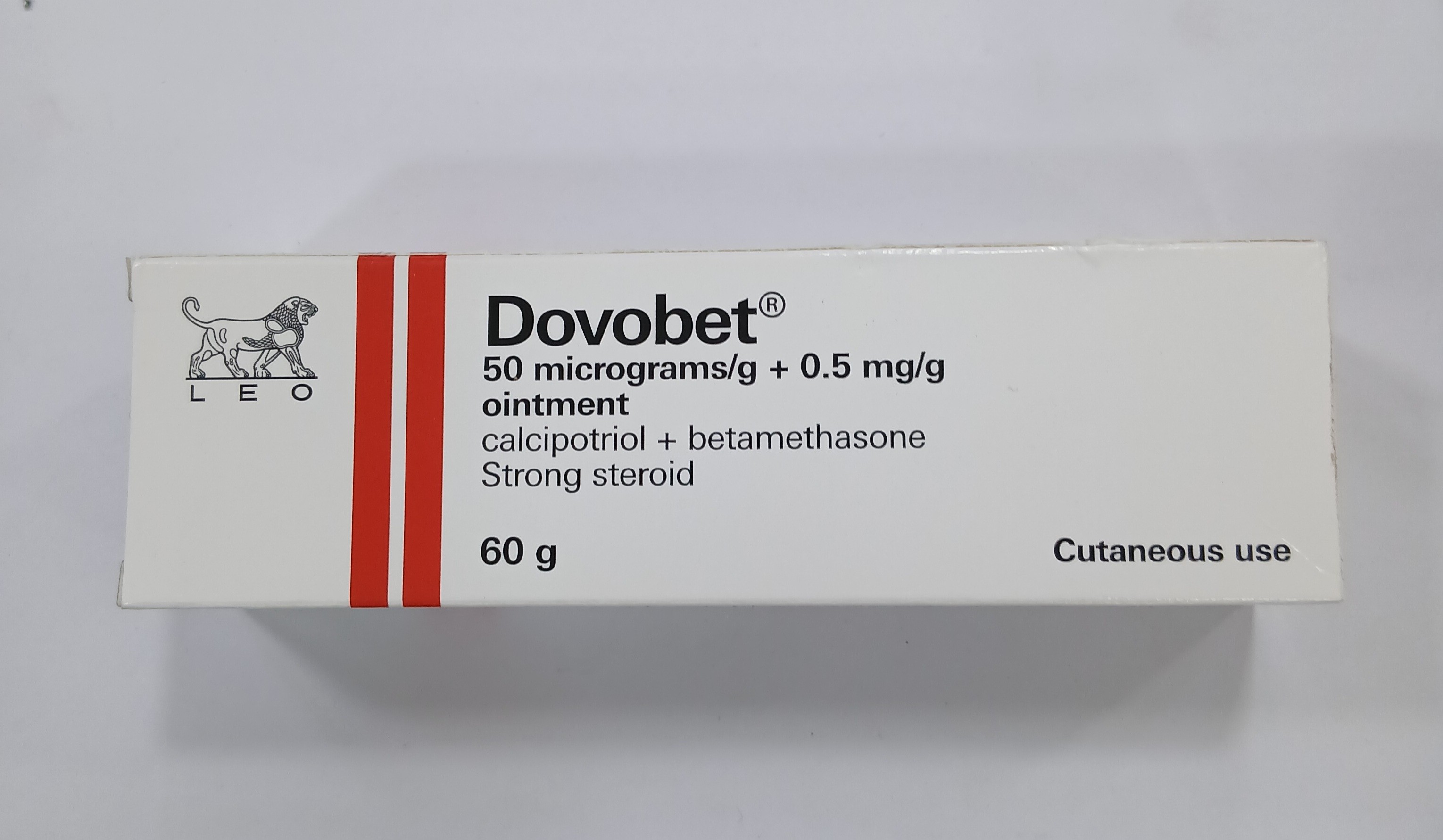 White cardboard box packaging displays a red and white label with printed text indicating a 60 gram ointment containing calcipotriol and betamethasone, marked for cutaneous use.