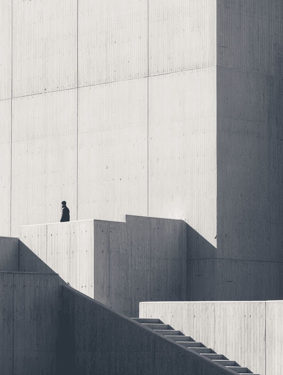 A lone person in dark clothing stands atop angular concrete platforms, flanked by stark stairs and towering light-gray walls that cast geometric shadows.