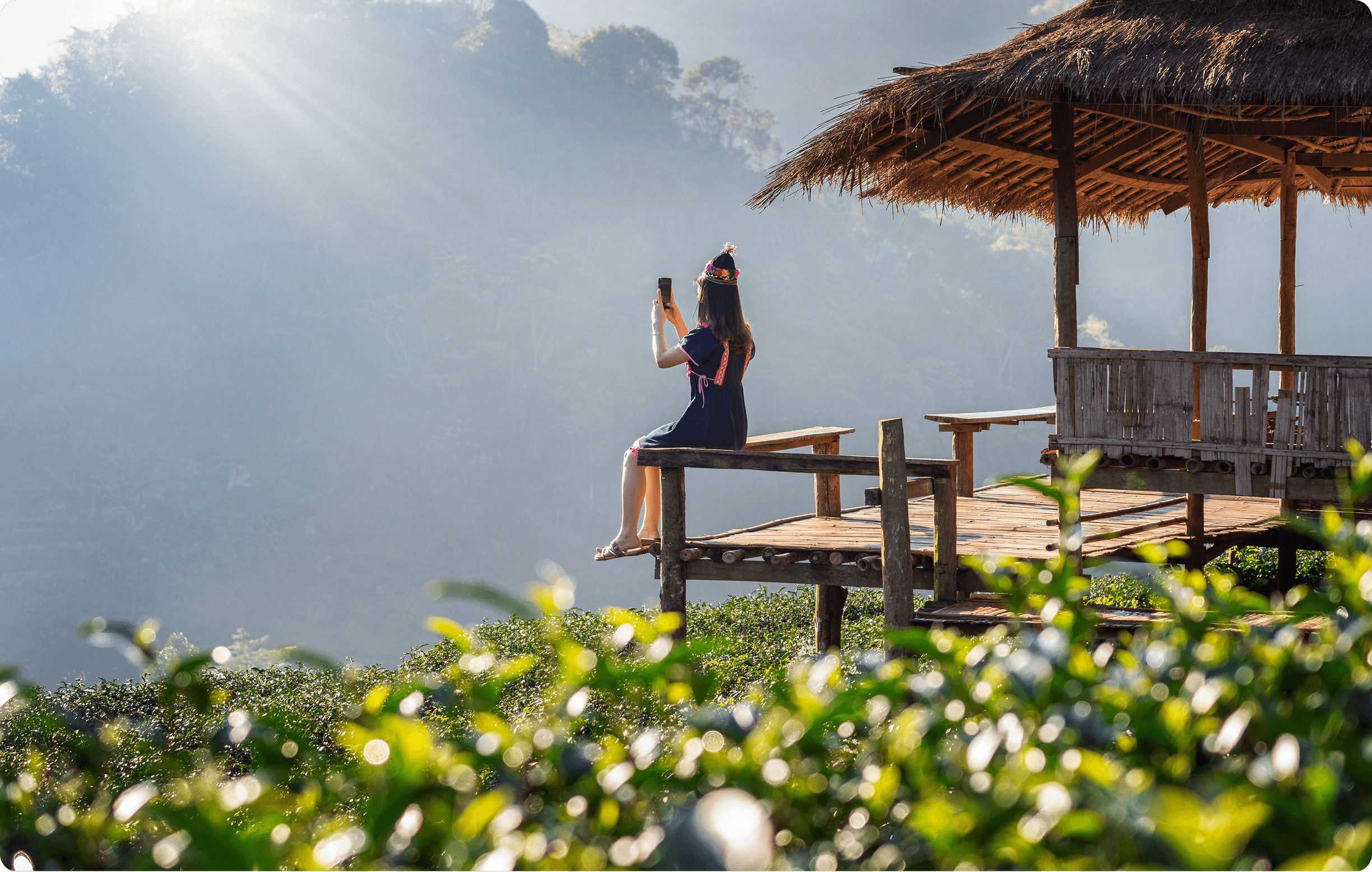 Woman sitting on a wooden hut deck taking a photo over a misty green valley.