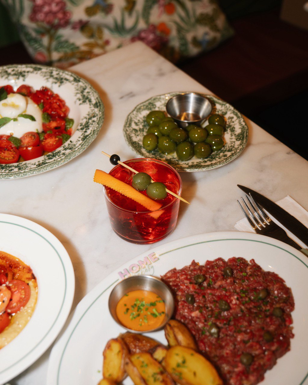 A marble table set with plates of vibrant dishes, including a colorful beet salad with tomatoes, a plate of seasoned green olives, and a platter featuring beef tartare adorned with capers, accompanied by roasted potatoes, alongside a bright red cocktail garnished with olives and a orange peel.